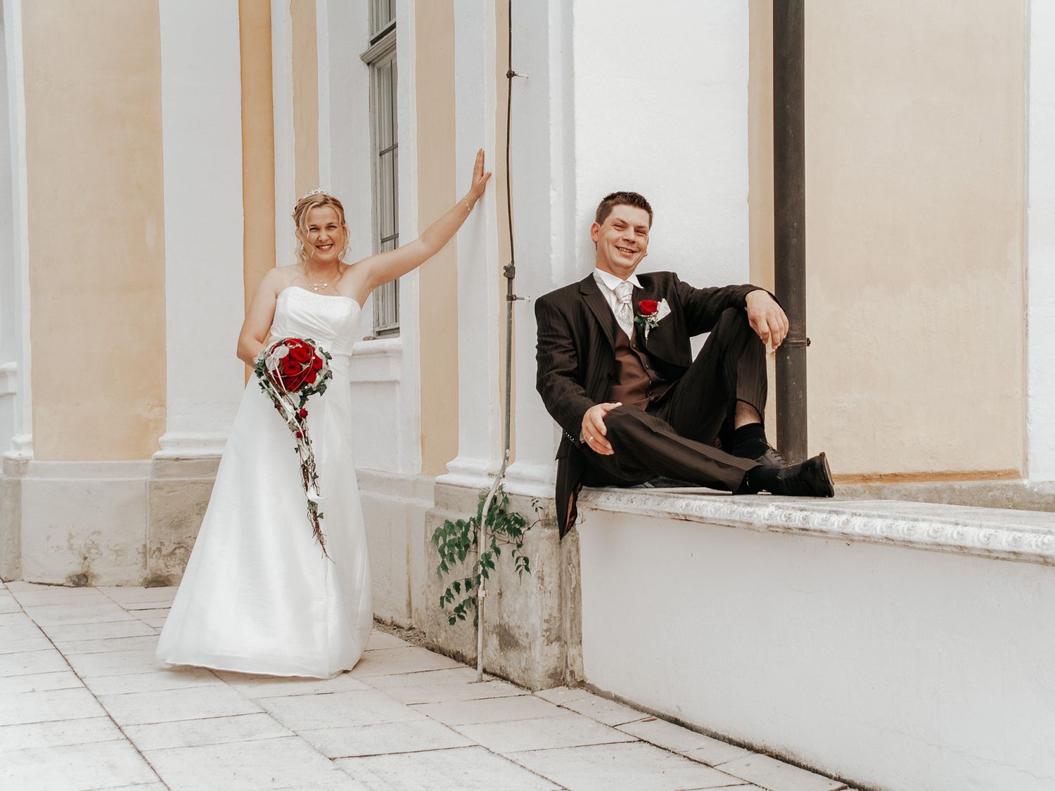 A bride and groom pose together in a light-filled corridor with white pillars and elegant architecture.