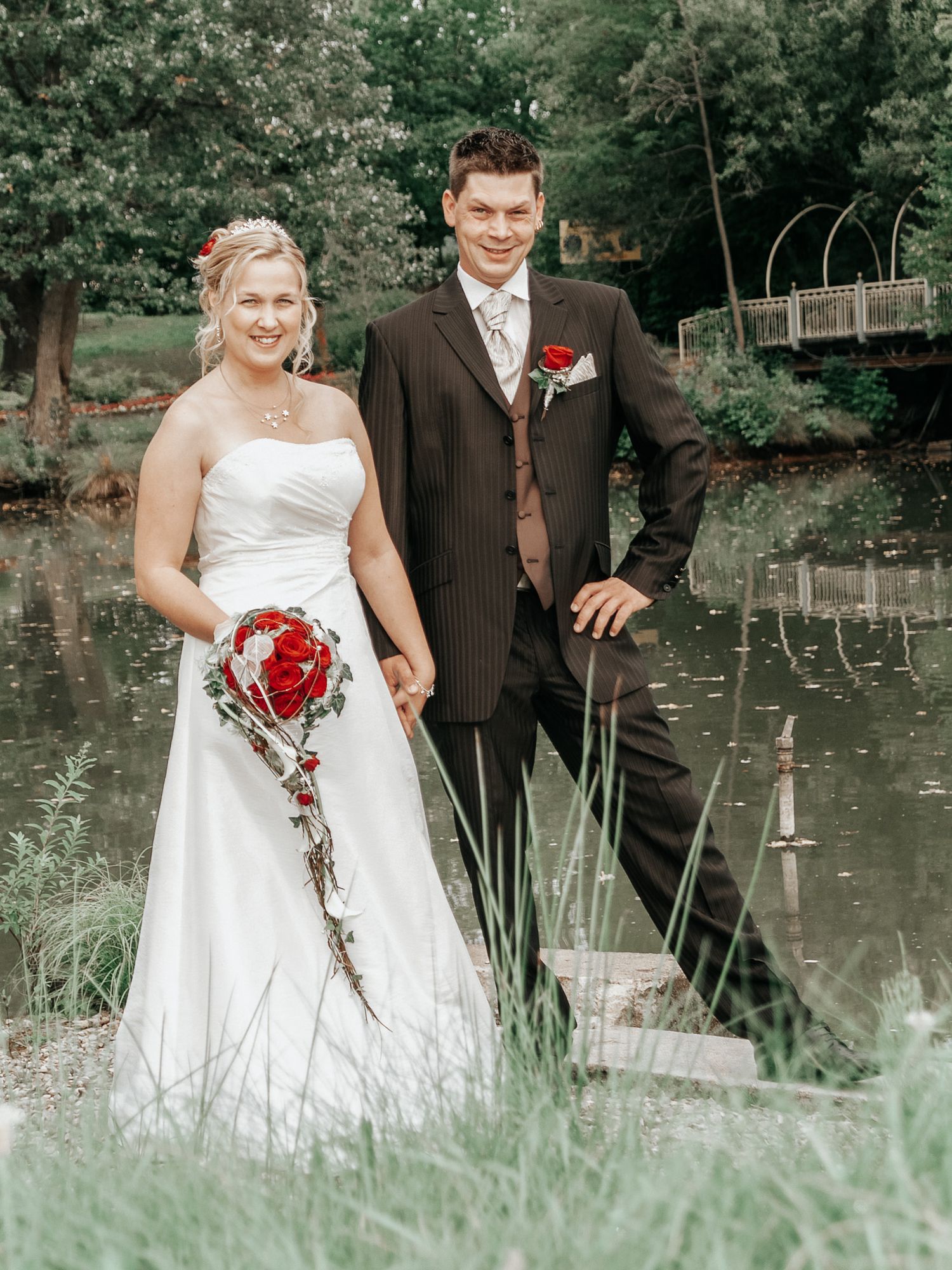 A wedding couple poses by a tranquil pond surrounded by greenery, with the bride holding a cascading red rose bouquet.