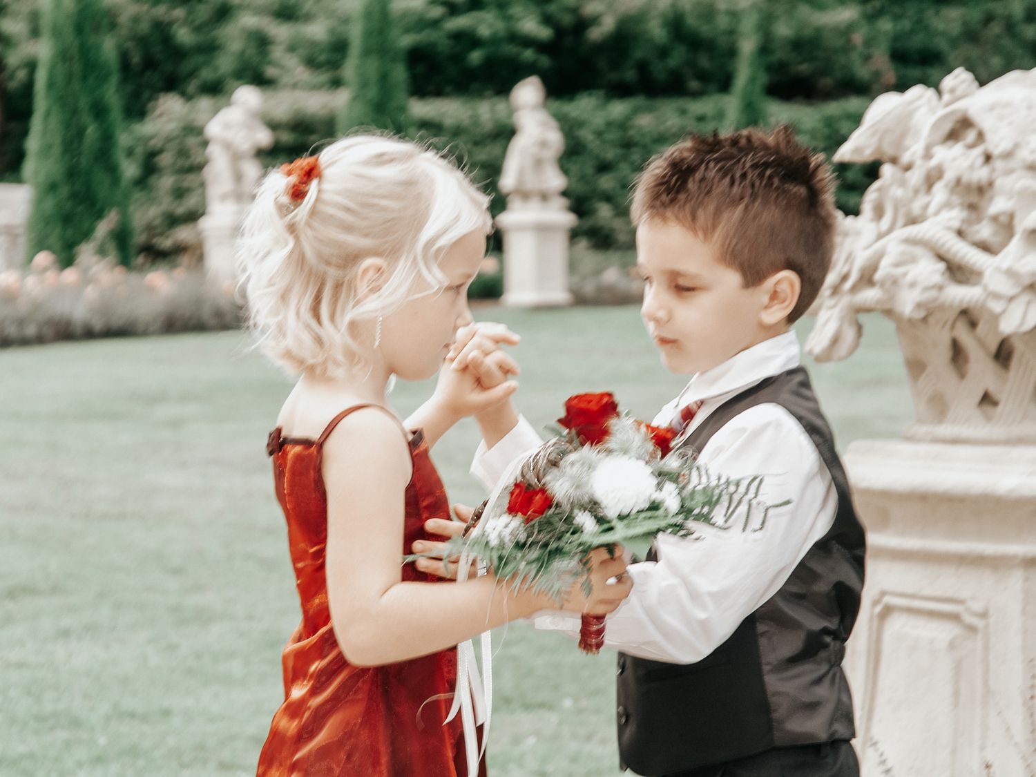 A couple in formal attire shares a romantic moment in a garden setting with ornate stone sculptures.