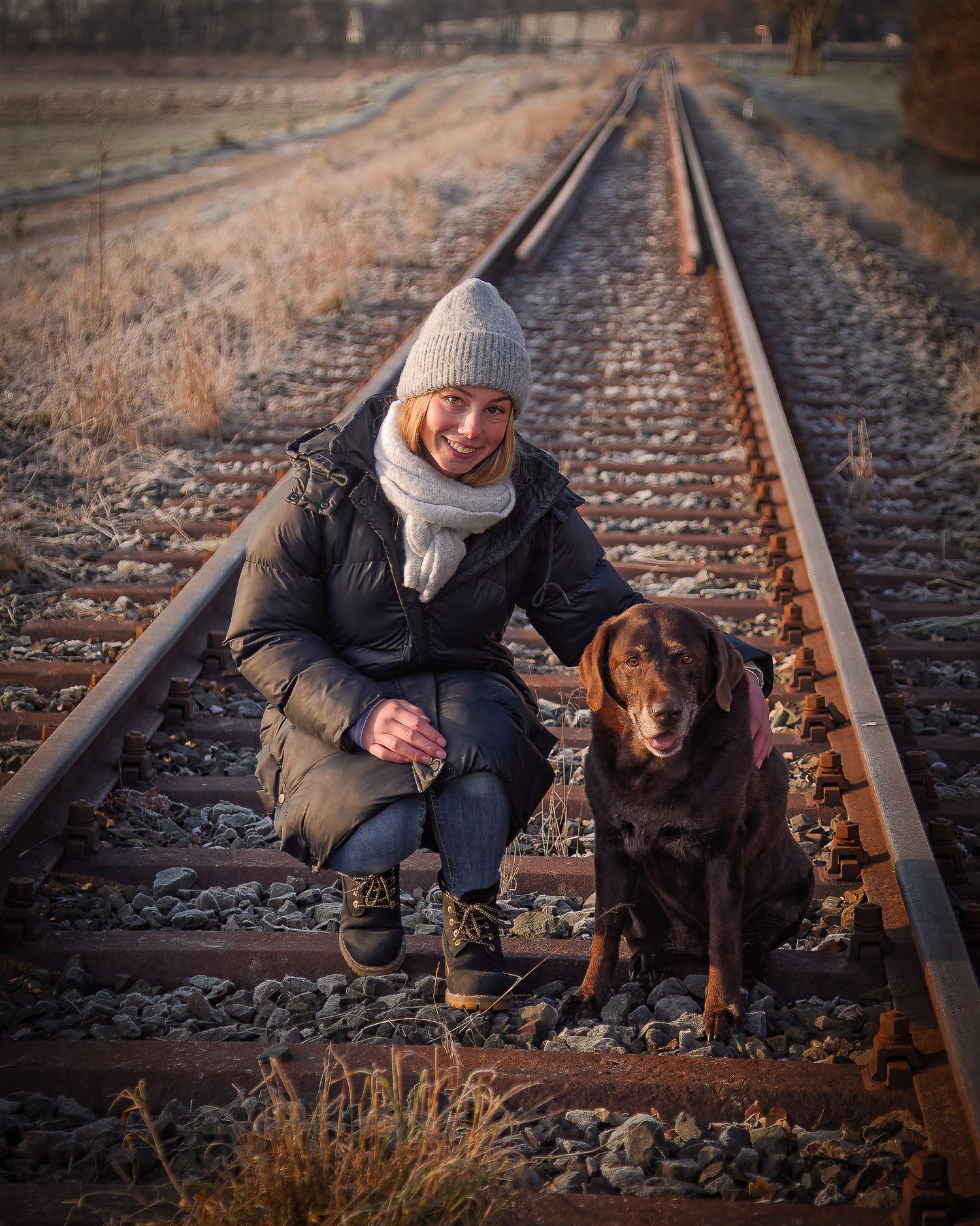 Eine Person in Winterkleidung sitzt mit ihrem Hund bei Sonnenuntergang auf Bahngleisen hier fährt auch die Dampfbahn Fränkische Schweiz.