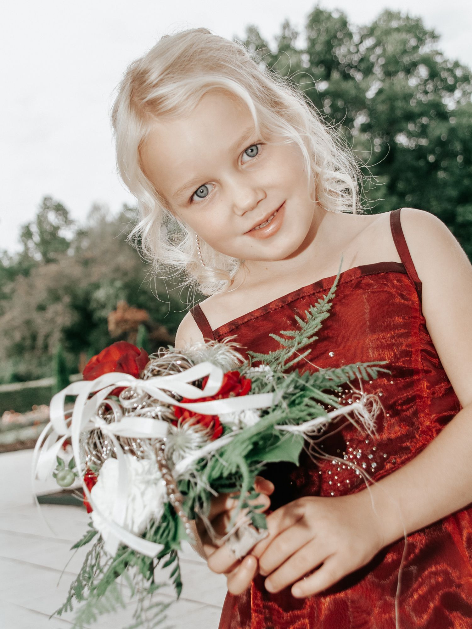 Child in red velvet dress holds winter greenery bouquet with white ribbons against outdoor background.