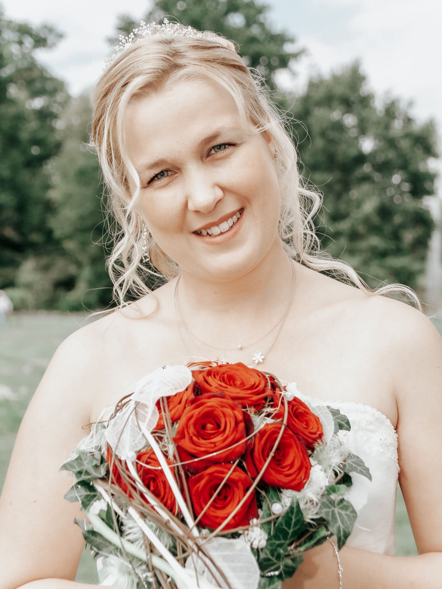 A joyful bride in a white dress holds a vibrant bouquet of red roses against a soft outdoor background.