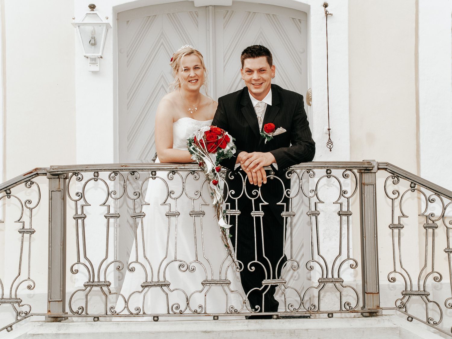 A wedding couple poses on an elegant white balcony with ornate iron railings for romantic portraits.