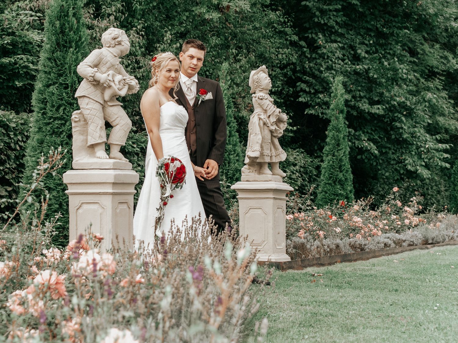 A couple in wedding attire poses together in a romantic garden setting with stone pillars and blooming roses.