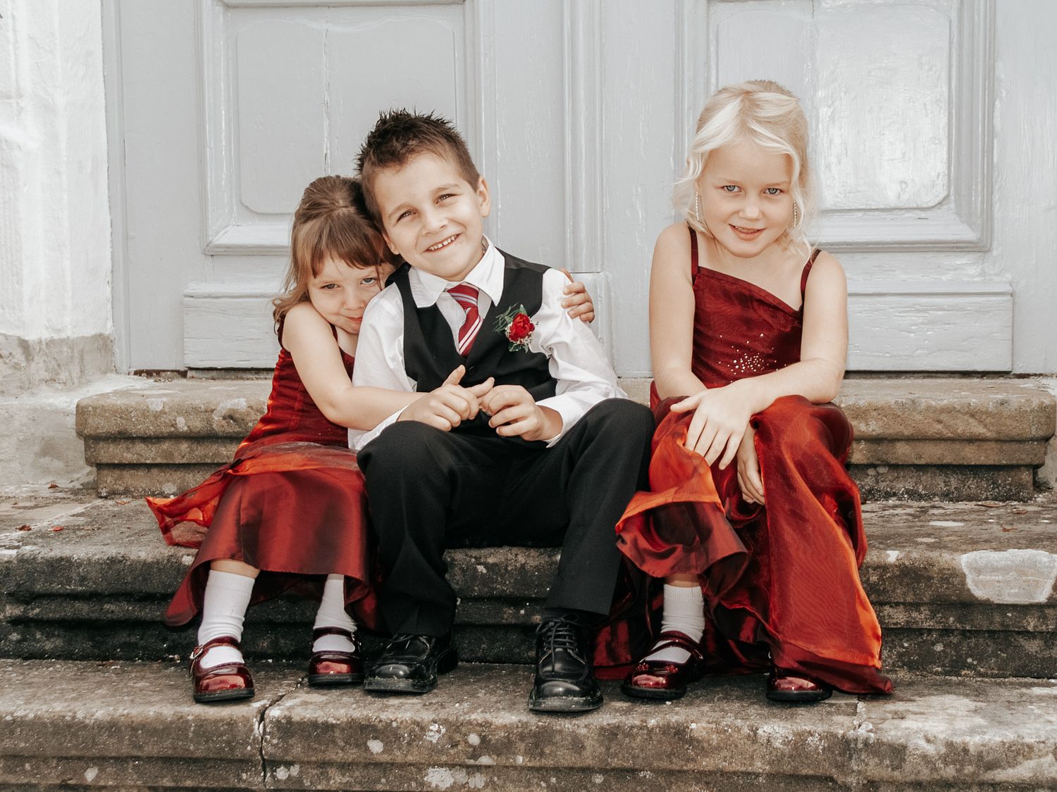 Children in formal attire sit playfully on stone steps, wearing red and black outfits.