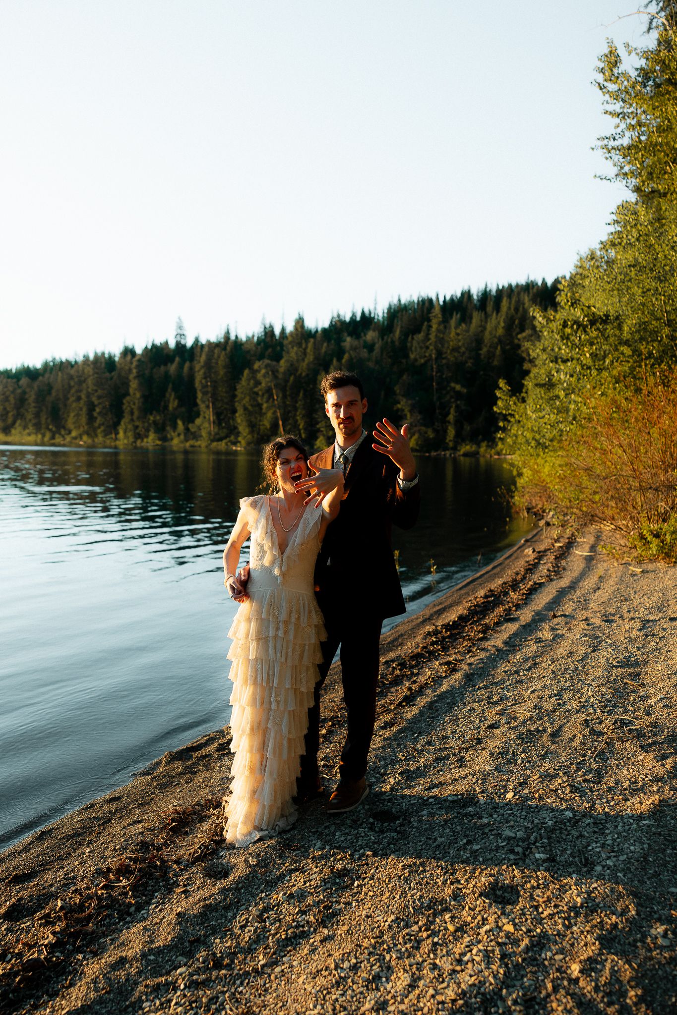 Couple in formal attire posing on a lakeside gravel shore surrounded by evergreen forest at golden hour.