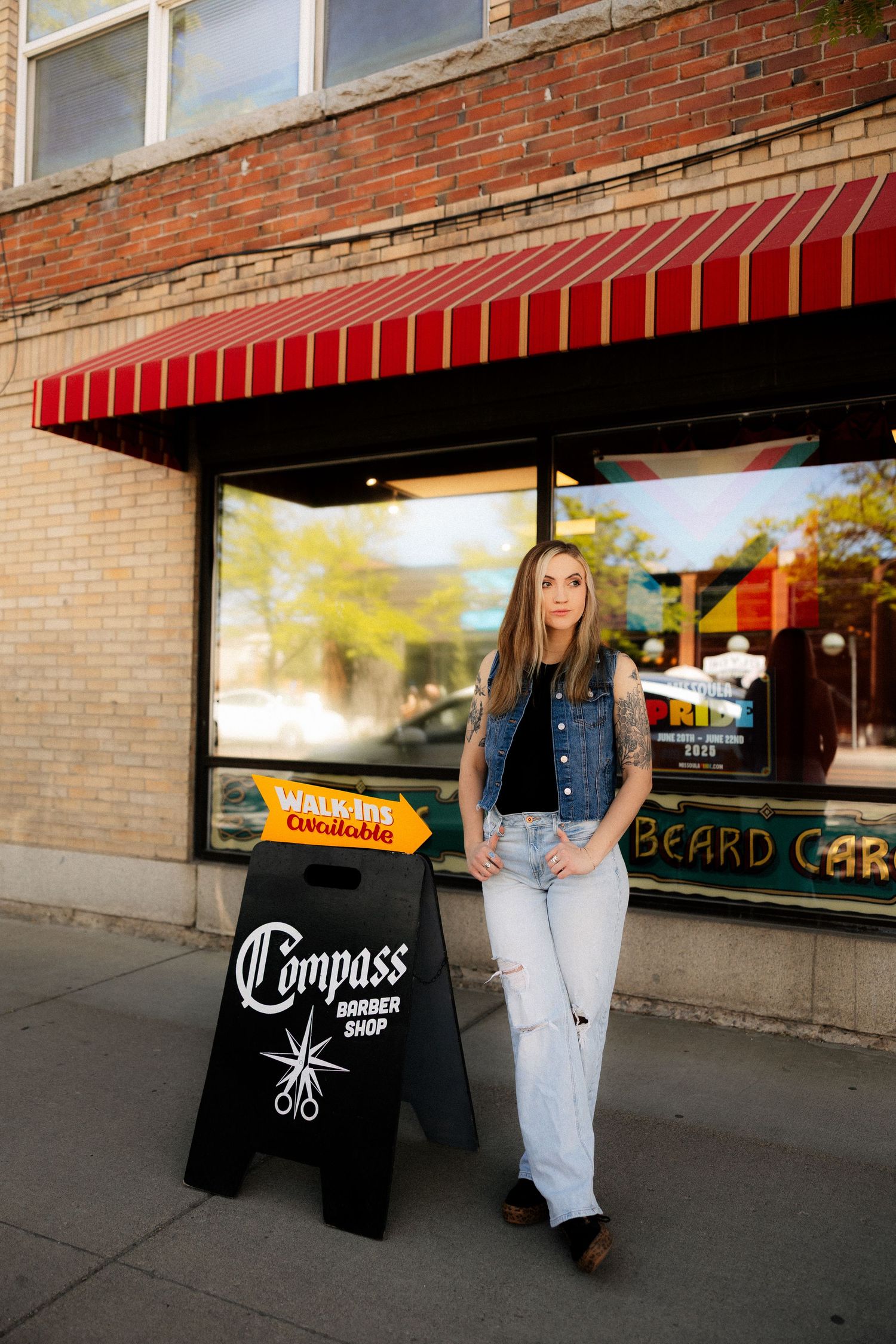 Young woman in a denim vest and ripped jeans posing beside a Compass Barber Shop sandwich board sign.