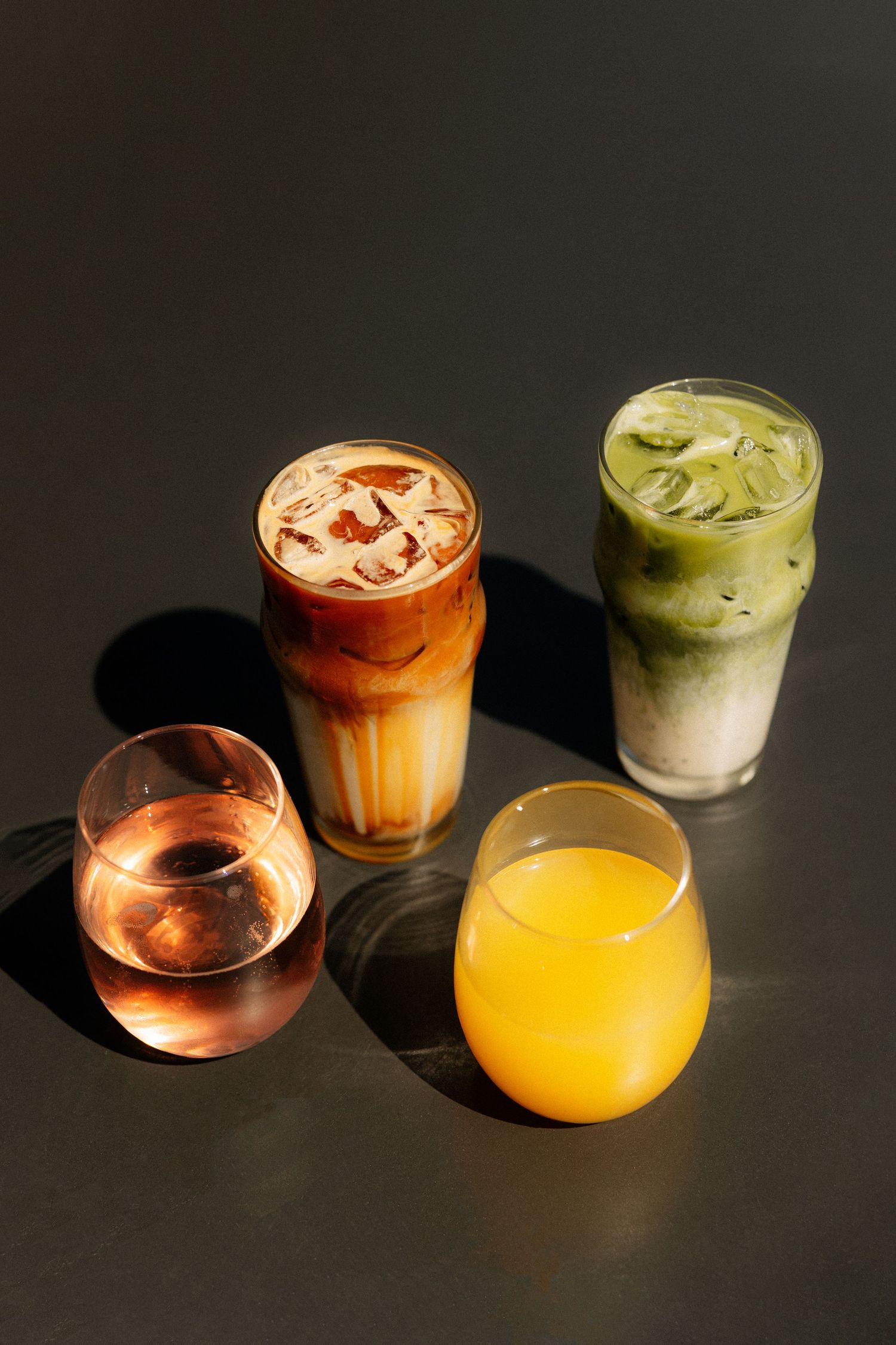 Four iced drinks on a dark surface including iced latte, matcha latte, orange juice, and rosé.