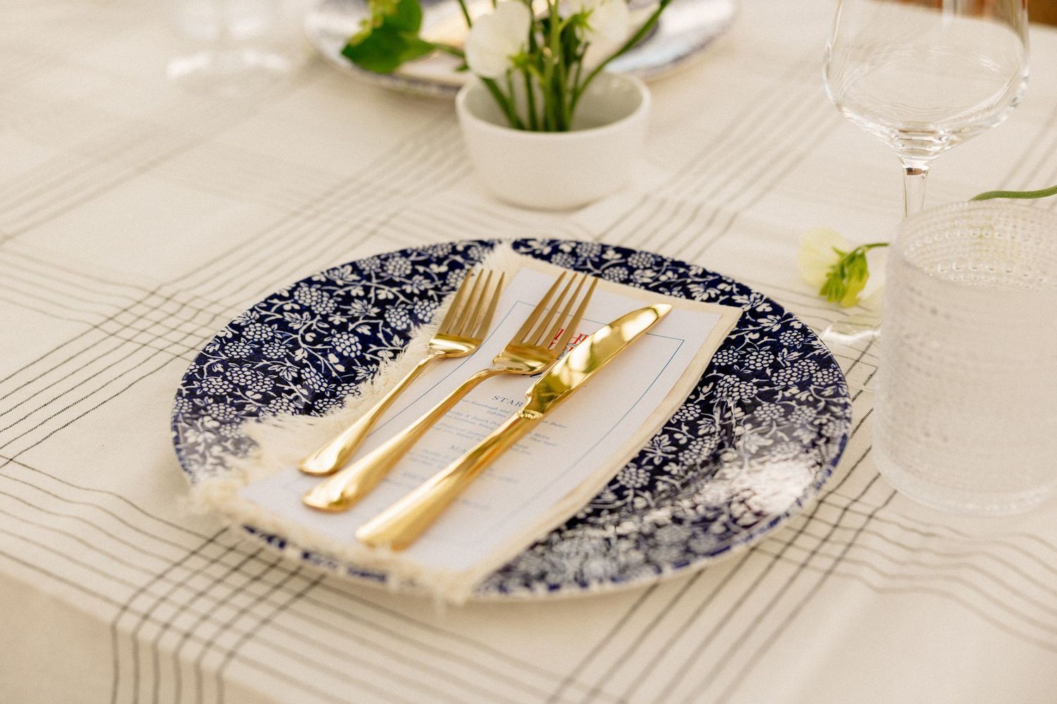 Elegant place setting with blue floral plate, gold cutlery, and white napkin on a striped tablecloth.
