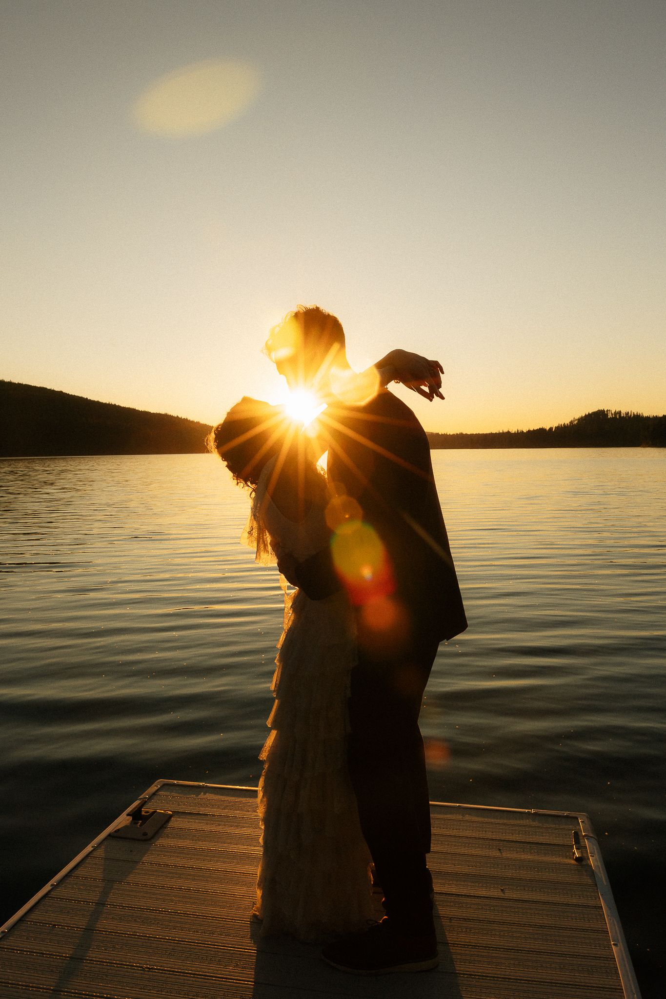 Silhouette of a couple kissing on a dock at golden sunset over a calm lake with lens flare.