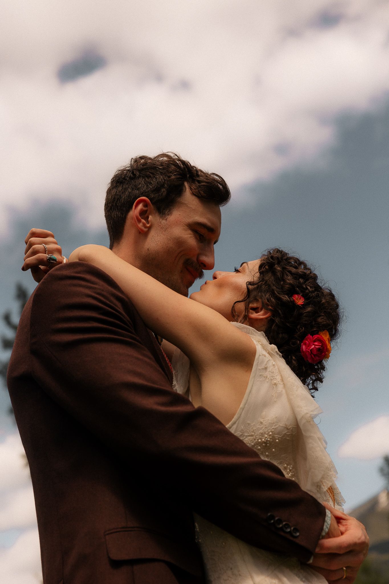 A bride and groom share a romantic kiss outdoors, dressed in a white gown and brown suit, with a cloudy sky backdrop.