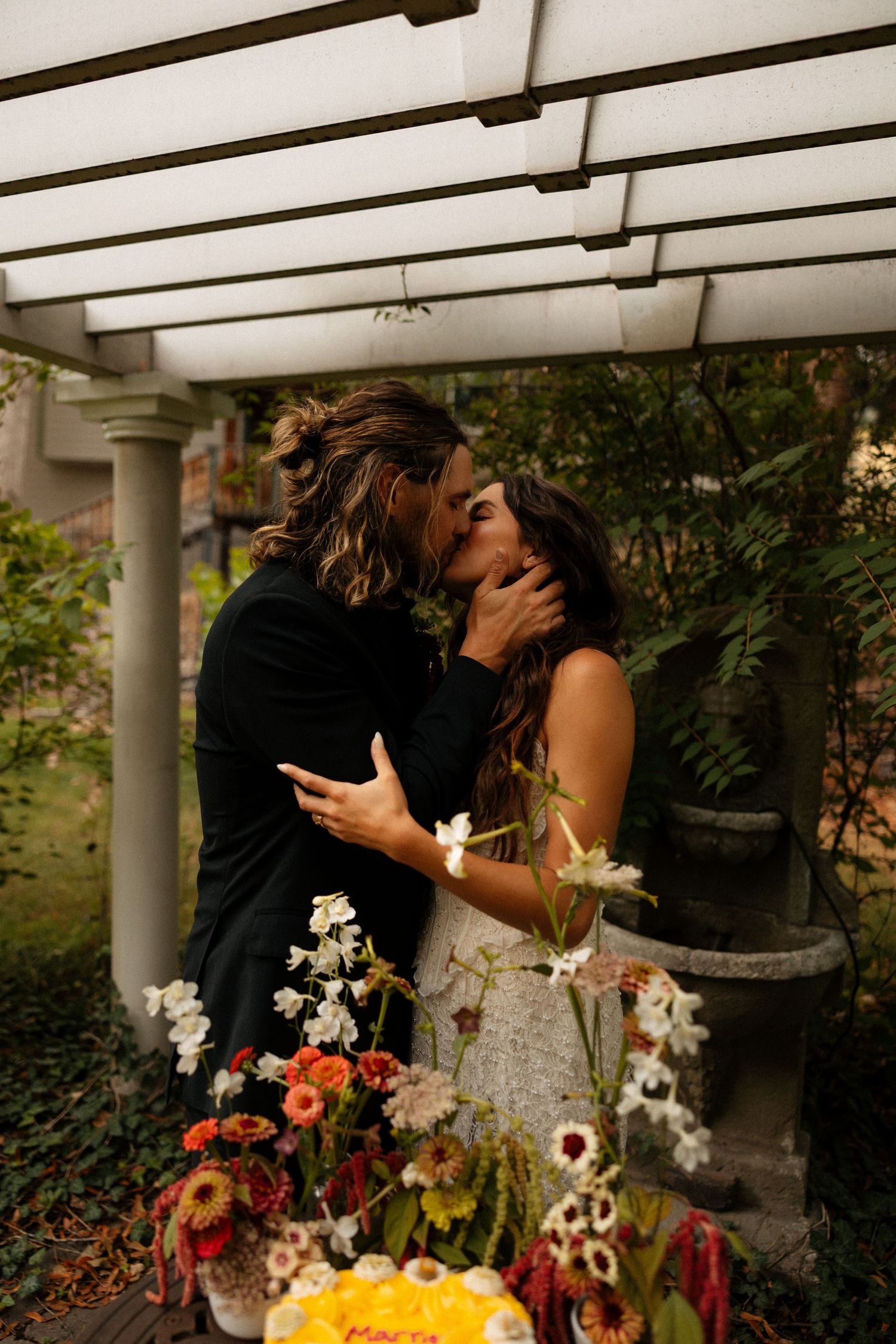 Couple sharing a romantic kiss under a pergola surrounded by colorful wedding flowers.