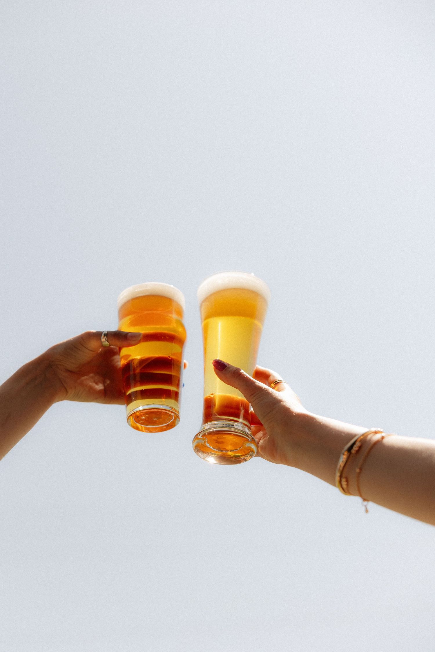Two hands clinking glasses of craft beer against a bright sky in a celebratory toast.