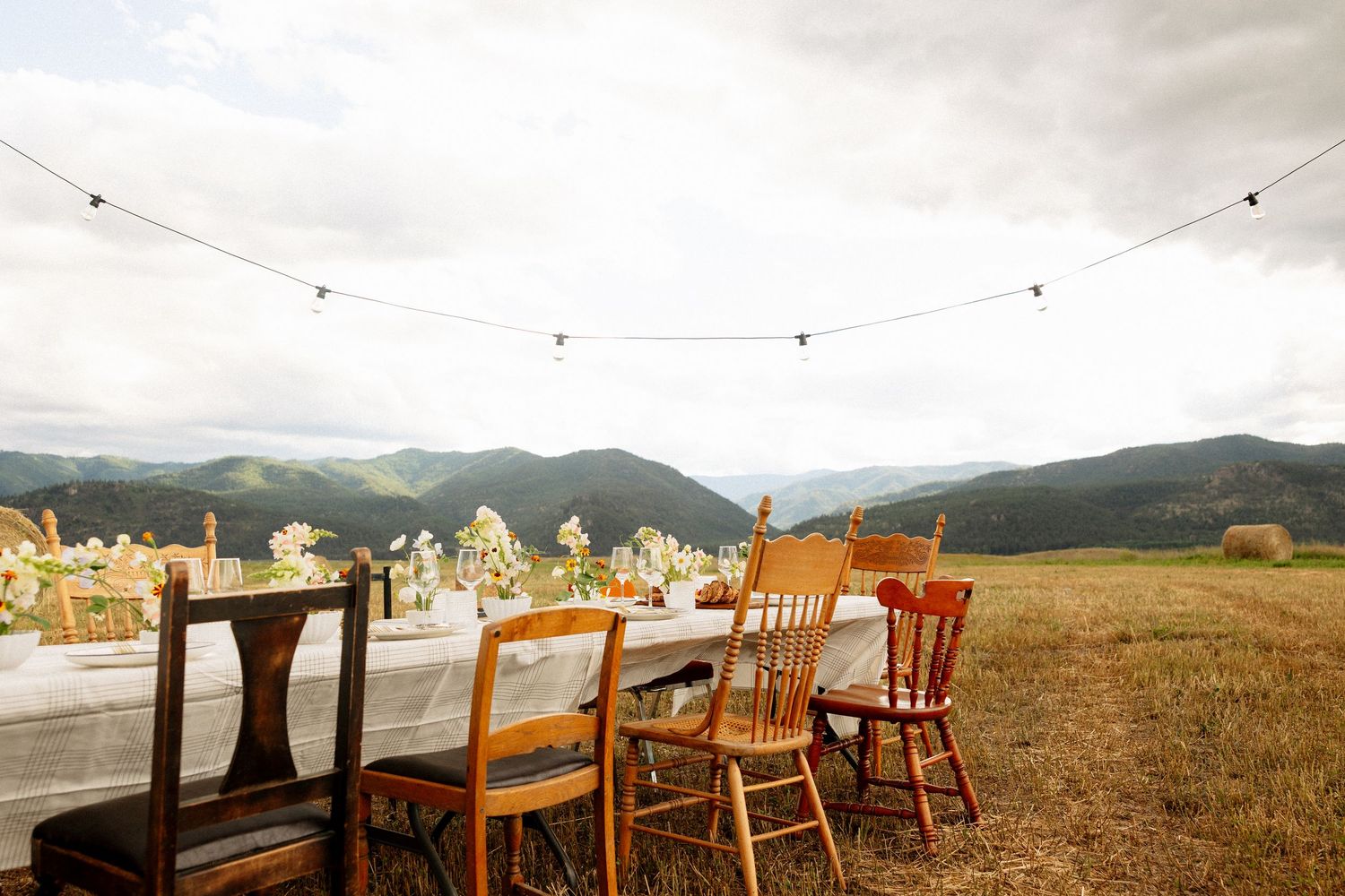 Outdoor dining table with wooden chairs set in a mountain meadow under string lights with scenic valley views.
