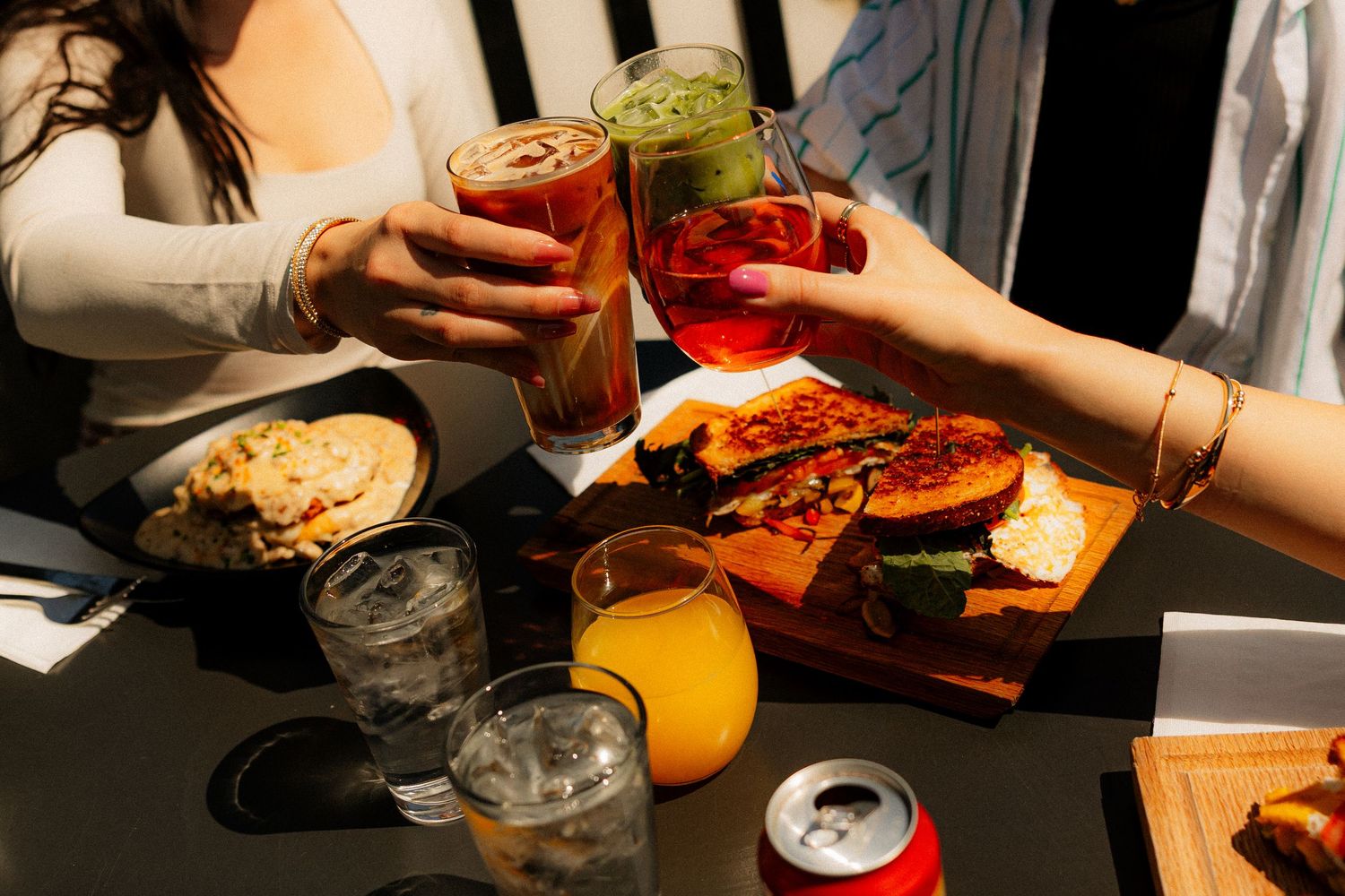 Friends toasting with colorful drinks over a table of food including sandwiches and snacks outdoors.