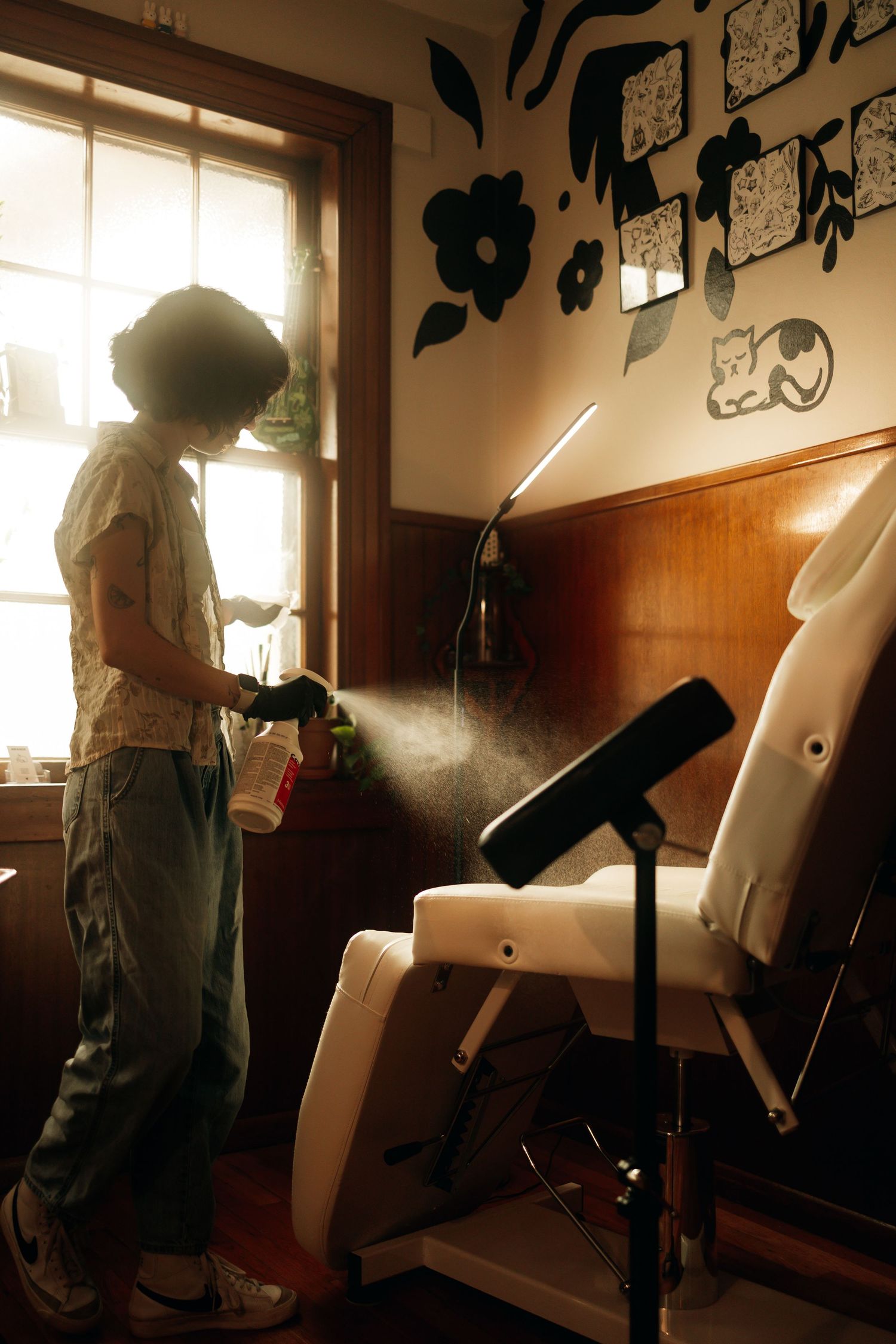 Person with afro sprays disinfectant on tattoo chair in sunlit vintage tattoo studio.