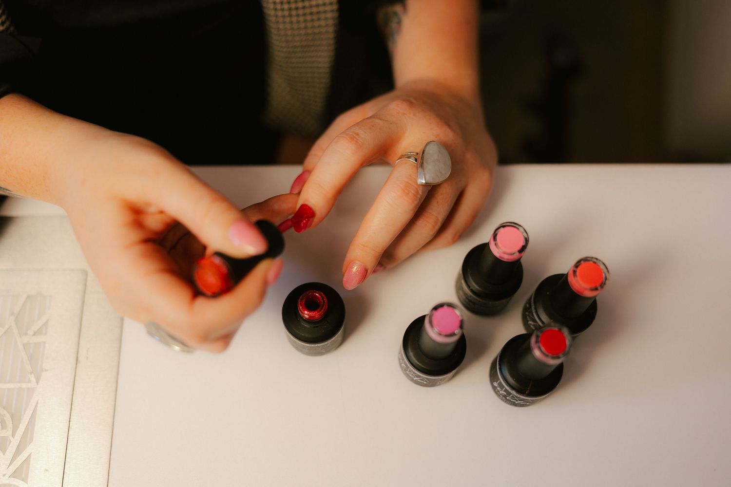 Close-up of hands with red nails selecting from a collection of pink and red lipsticks on a white surface.