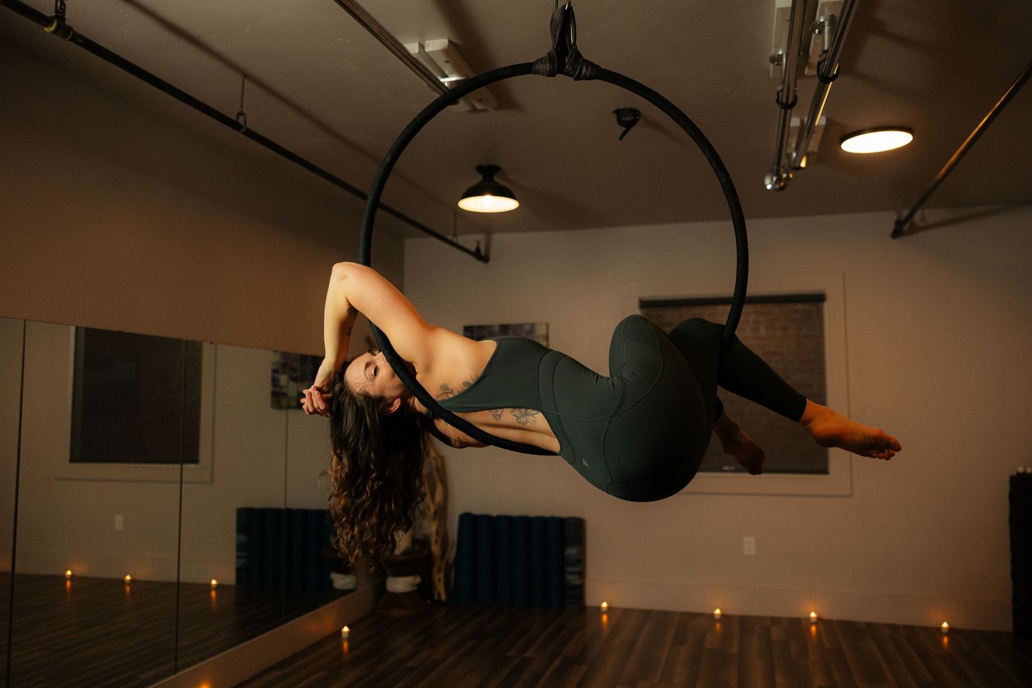 Female aerialist in green bodysuit performing an inverted pose on an aerial hoop in a candlelit studio.