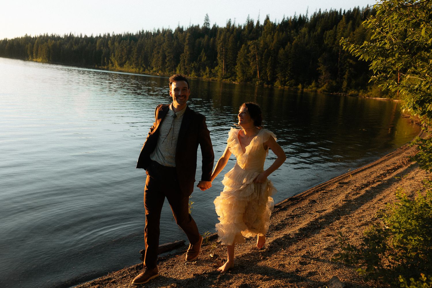 Bride and groom holding hands walking along a lakeside shore at golden hour with evergreen forest backdrop.