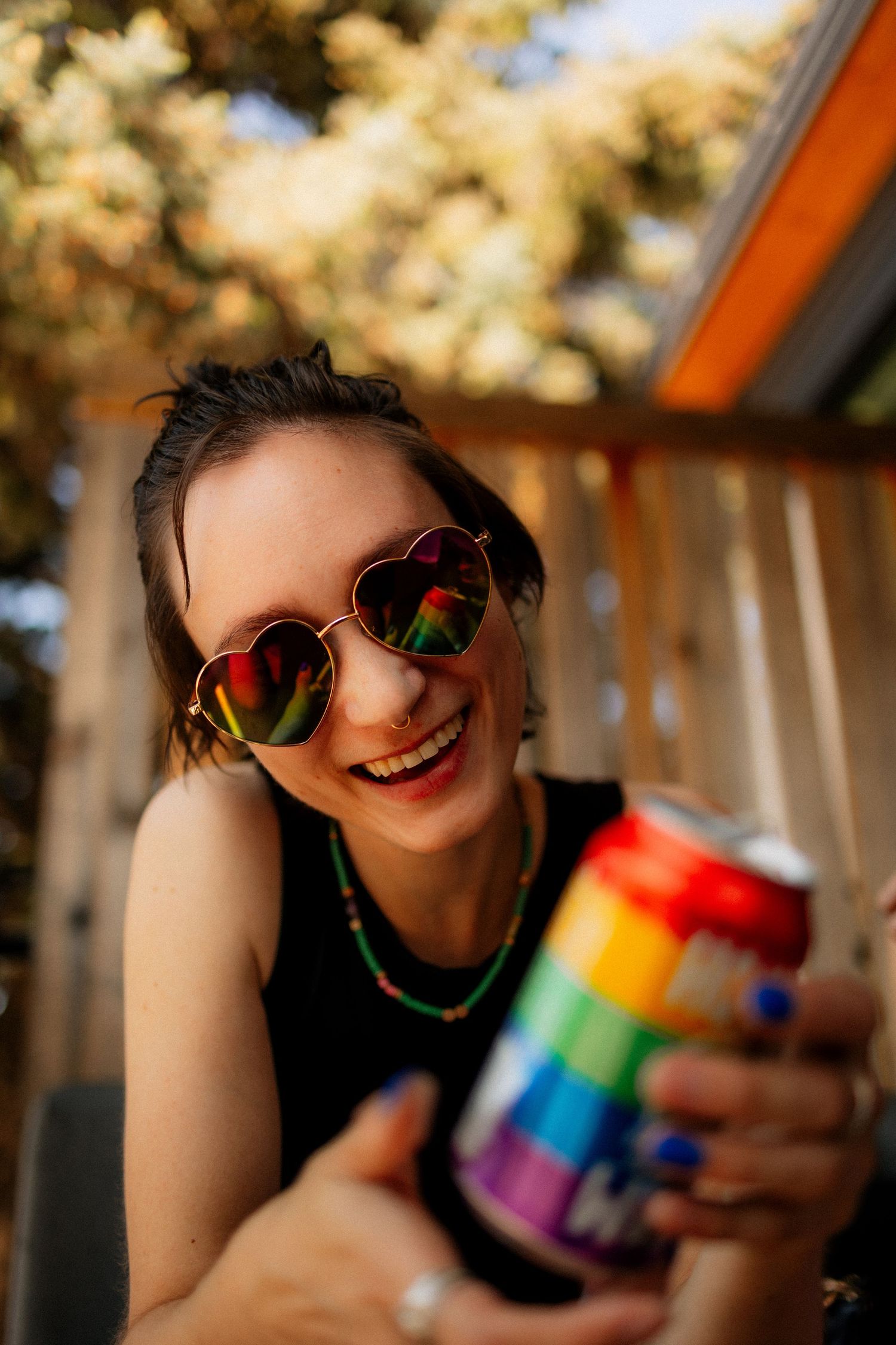 Smiling woman in heart-shaped sunglasses holding a rainbow Pride-themed beverage can outdoors.