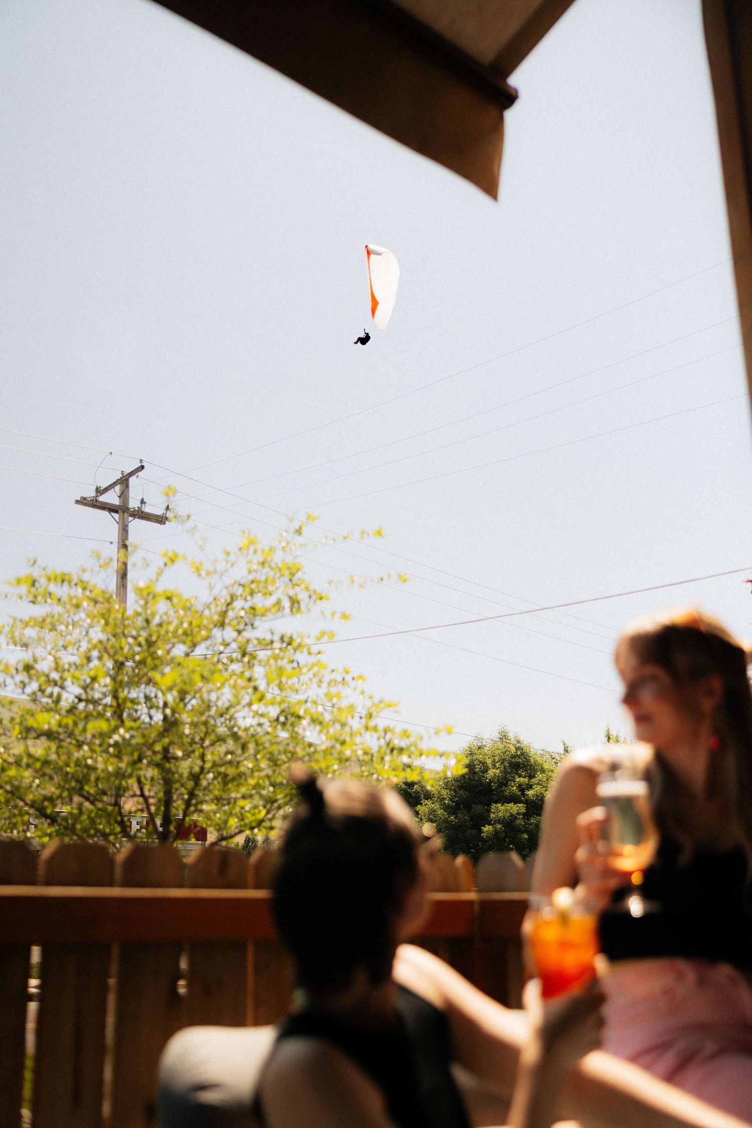 Two women enjoying drinks on a patio while a paraglider soars in the blue sky above.