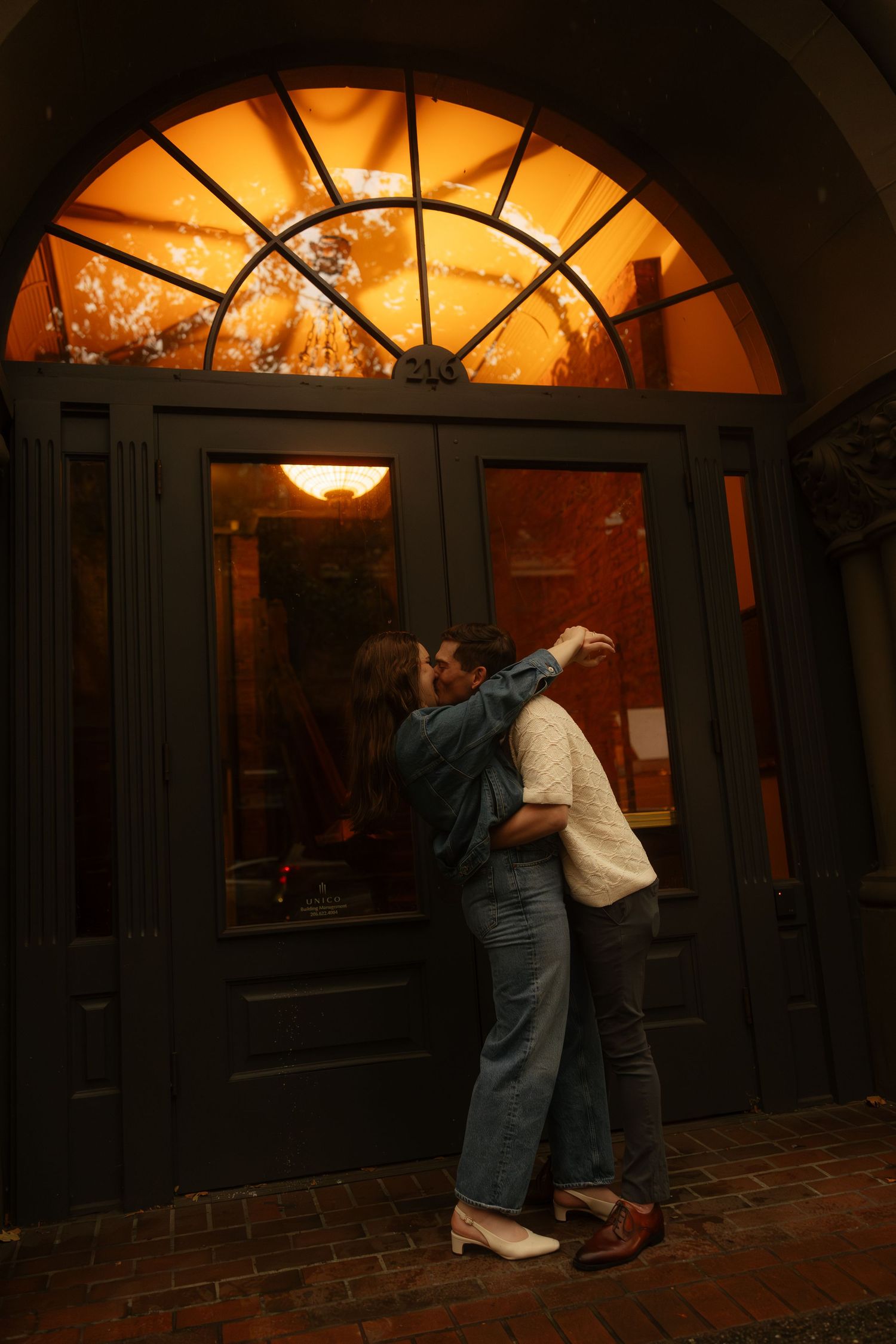 Couple sharing a kiss outside an elegant arched doorway glowing with warm amber light.
