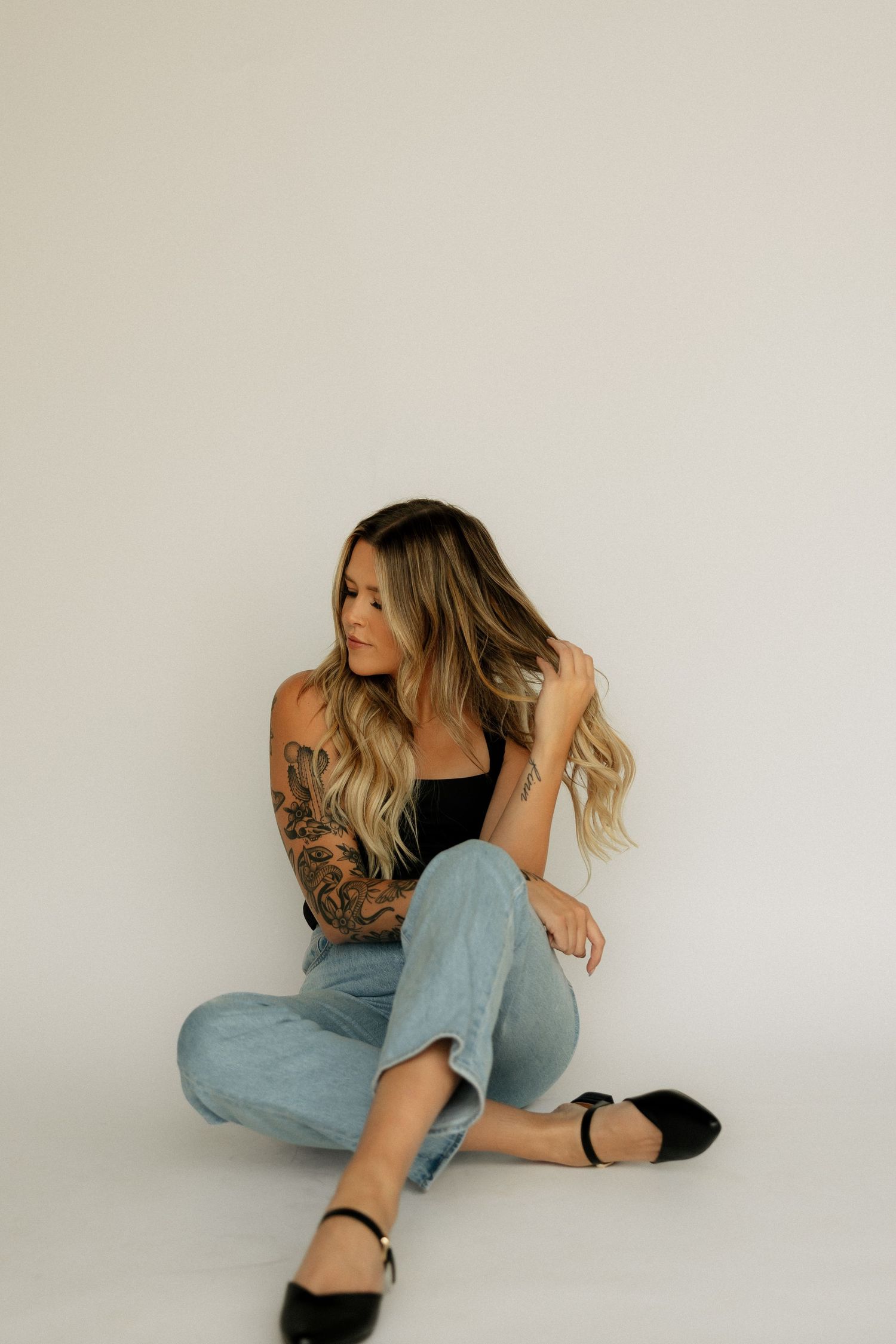 Tattooed blonde woman in black bodysuit and light jeans sitting casually on floor against white background.