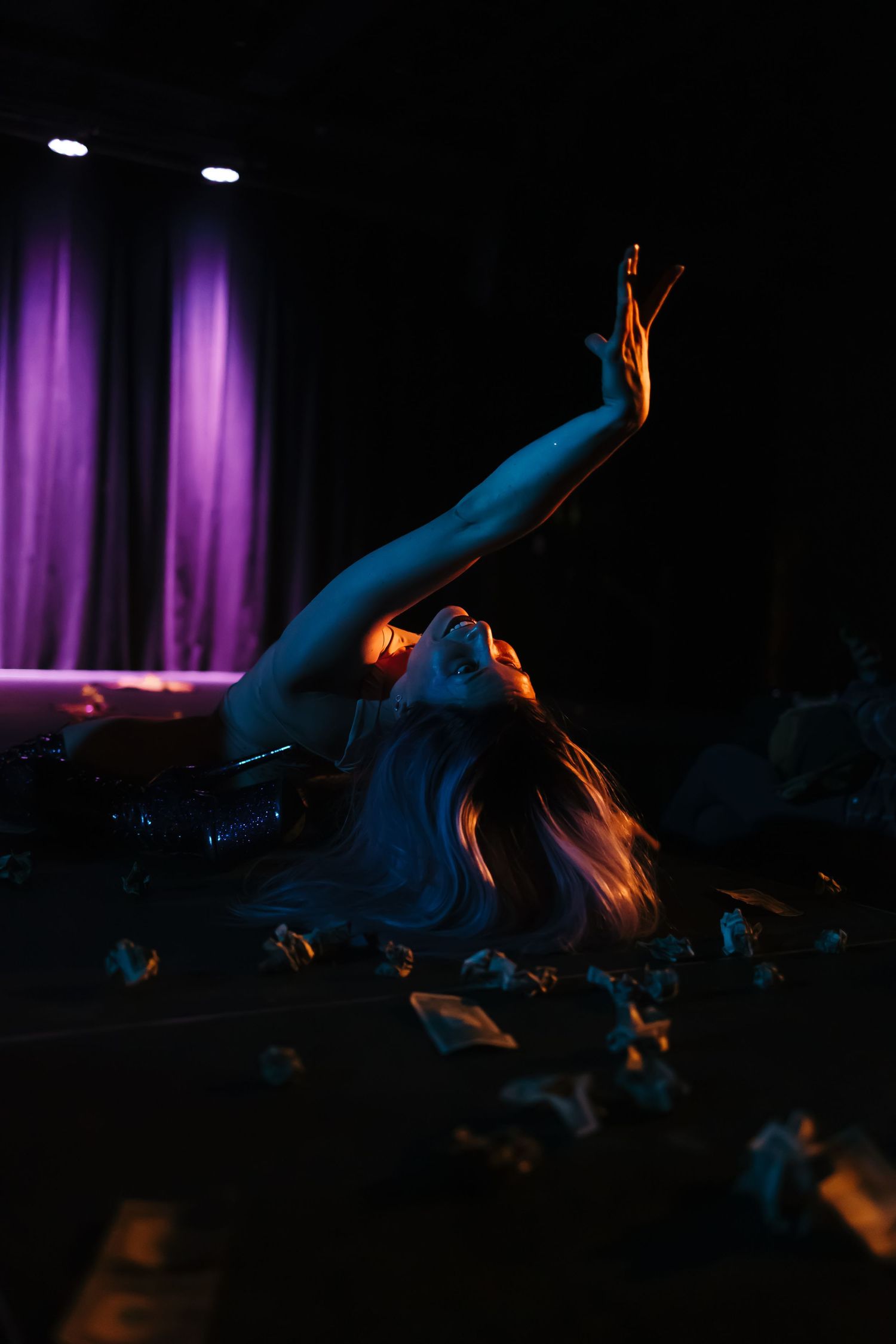 Dramatic stage performance with a woman lying on a dark floor reaching upward under purple and orange lighting.