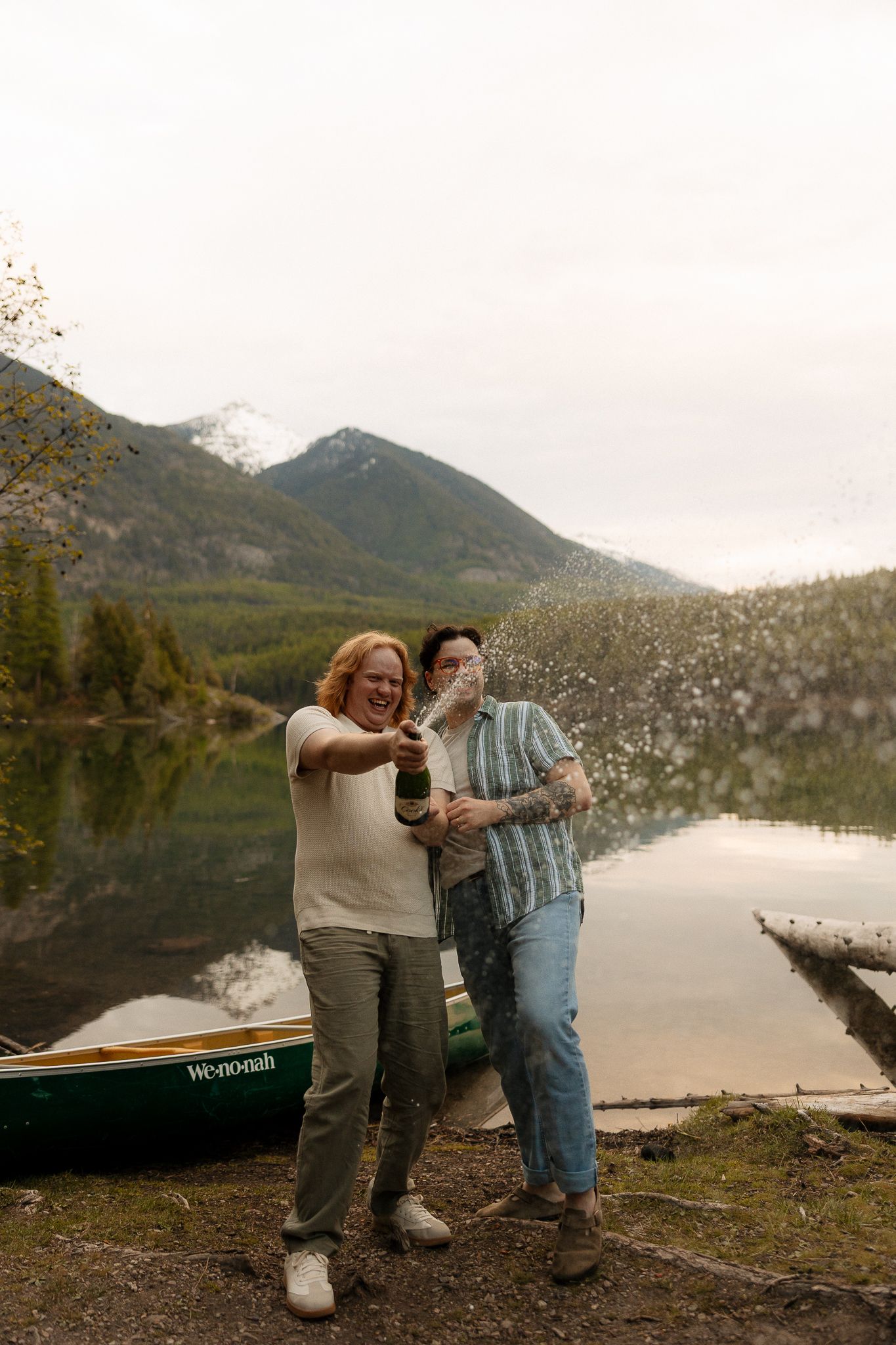 Couple celebrating by a mountain lake, popping champagne beside a green Wenonah canoe.