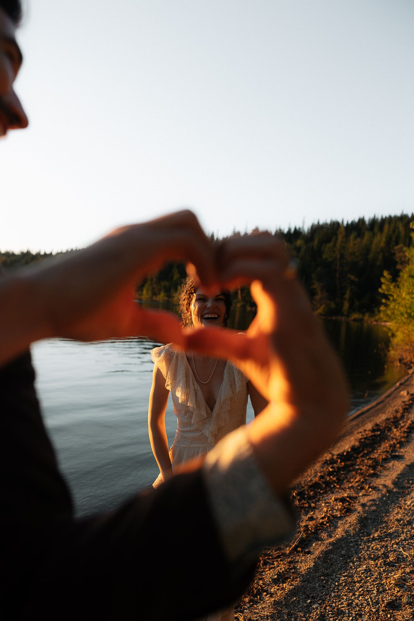 Couple making a heart shape with hands near a lake at golden hour, woman in white dress smiling in background.