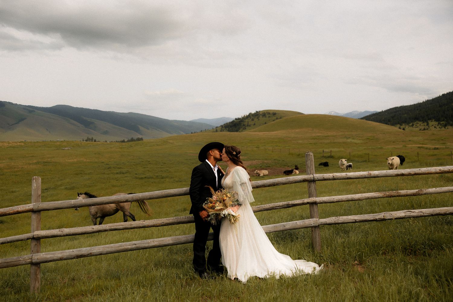 Bride and groom share a kiss leaning against a wooden fence with horses grazing in a sweeping green mountain valley in Montana
