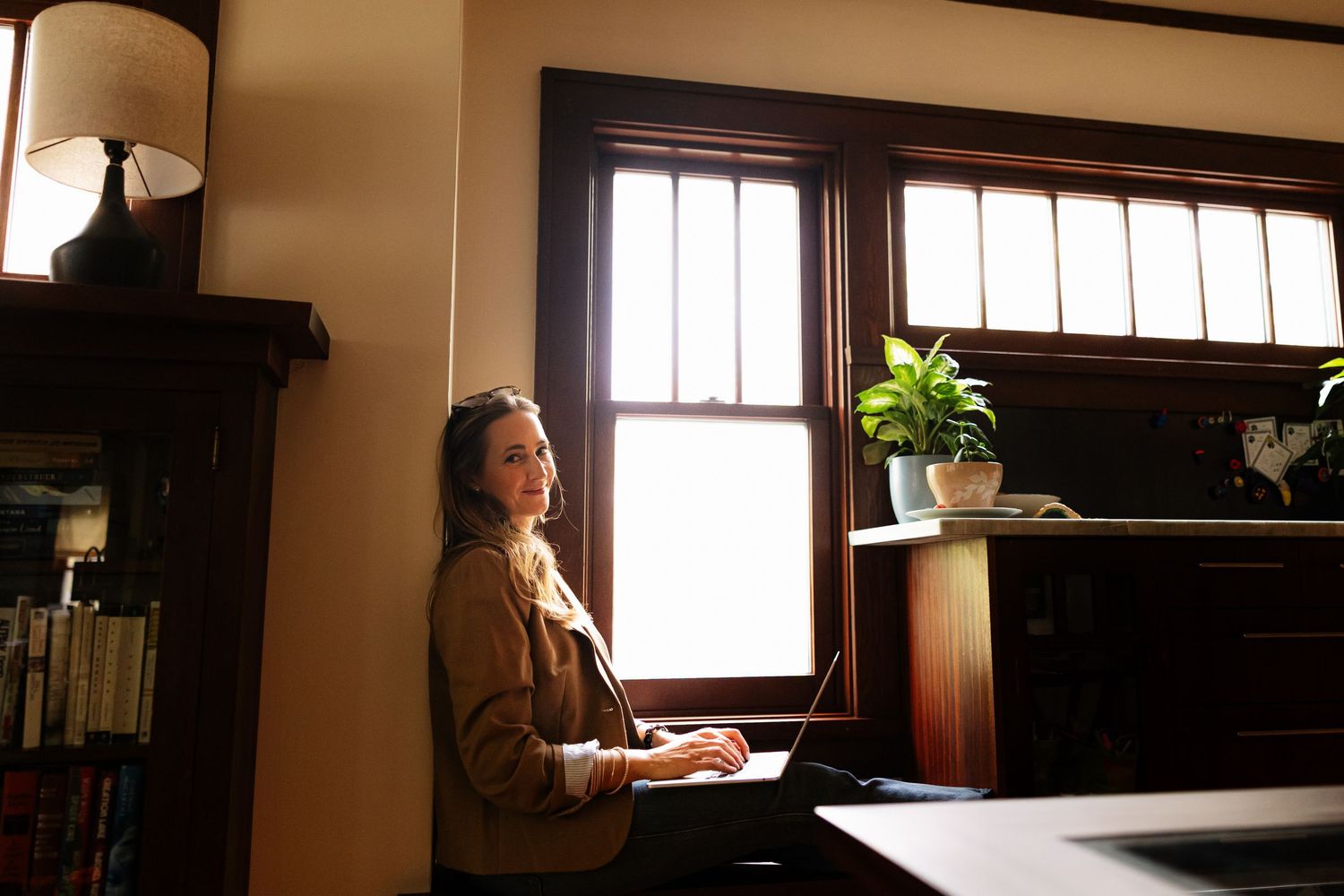 Woman working on laptop at desk by sunlit window in elegant wood-paneled home office.