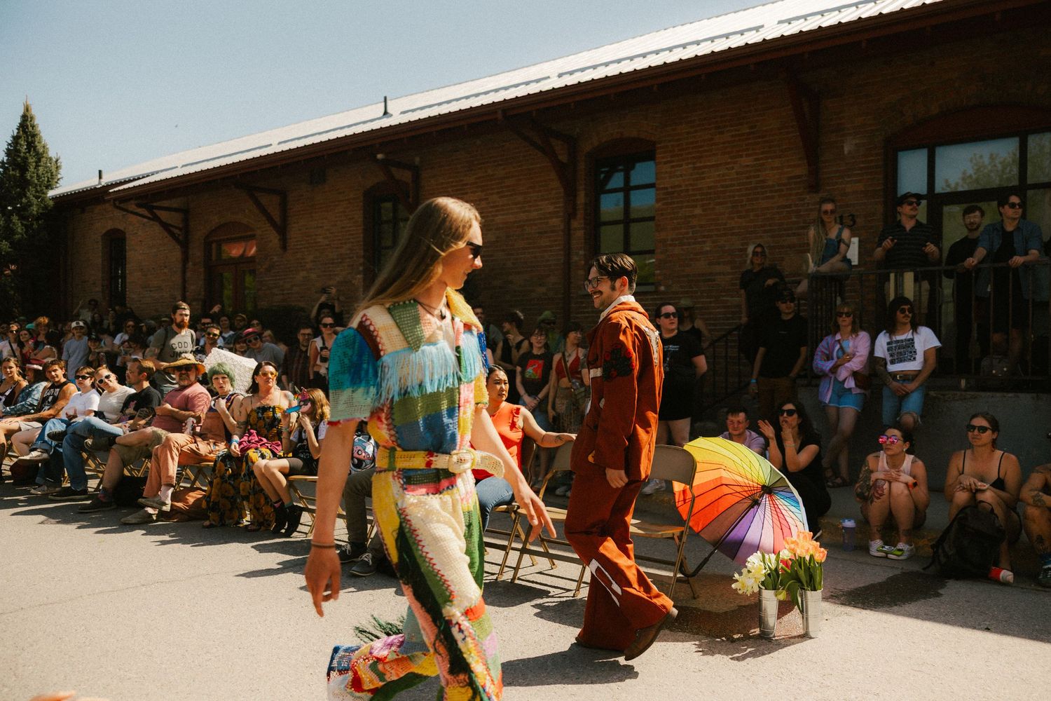 Models in colorful bohemian outfits walking an outdoor fashion runway before a large crowd of spectators.