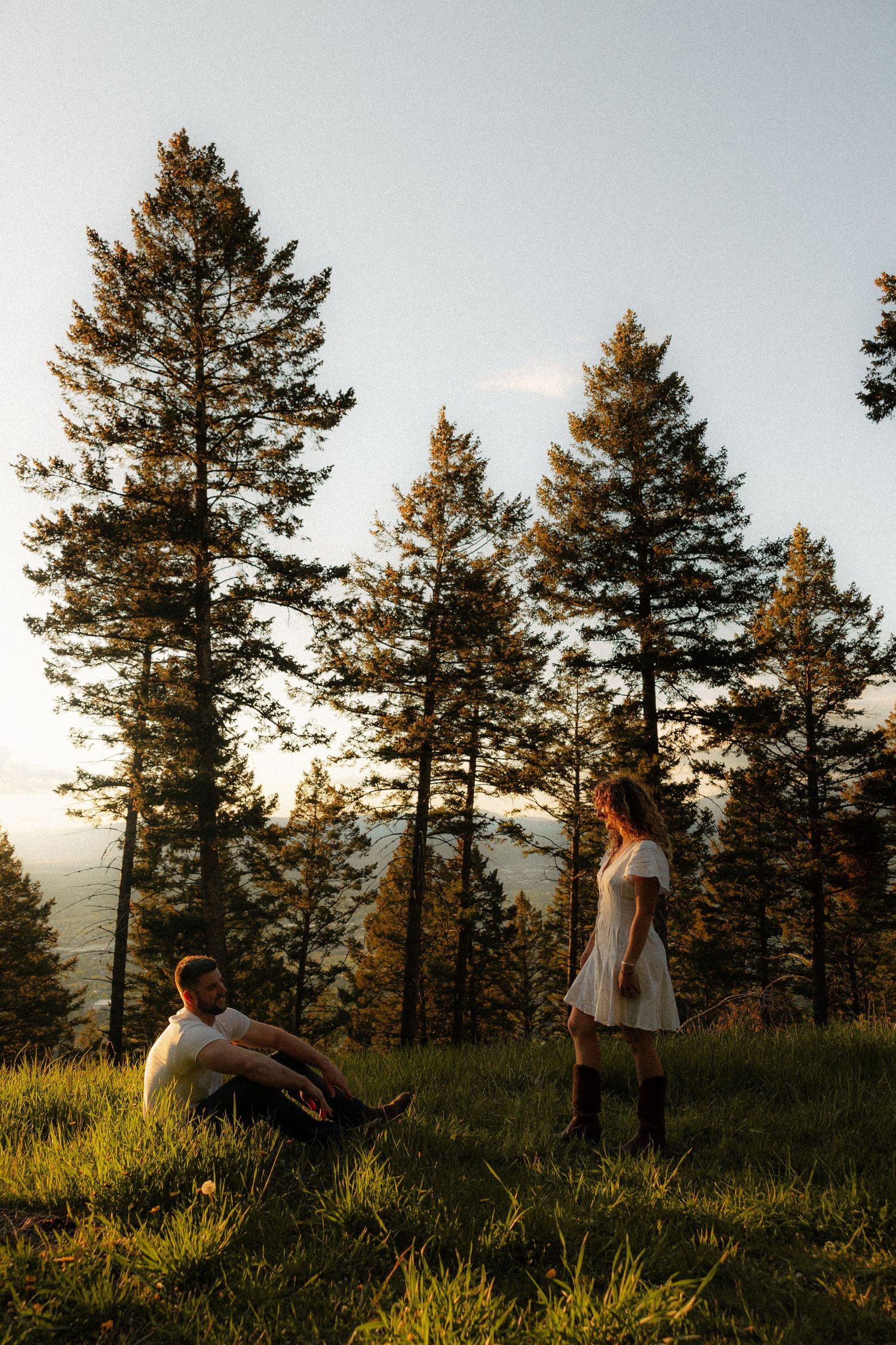 Couple enjoying a golden hour sunset moment on a grassy hillside among tall pine trees.