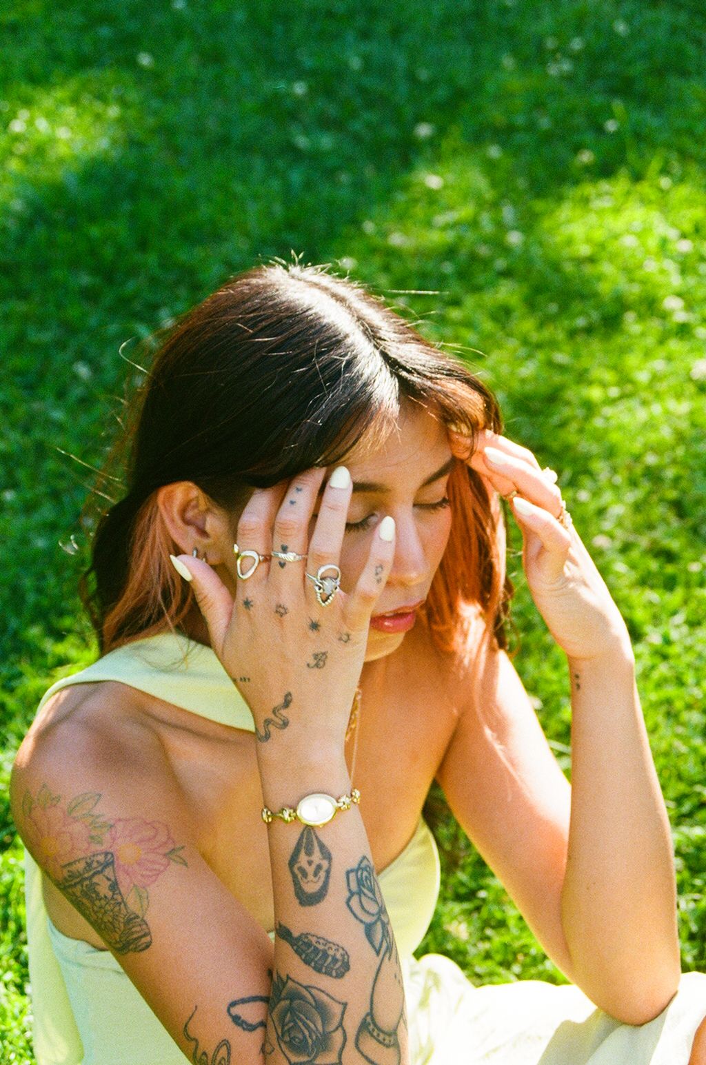 Tattooed woman in yellow dress sits on green grass, covering her face with hands adorned with gold rings and a bracelet.