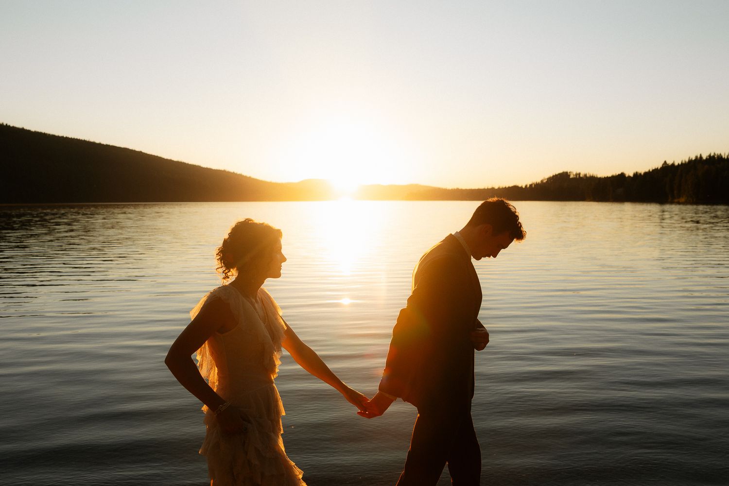 Couple holding hands silhouetted against golden sunset over calm lake waters.