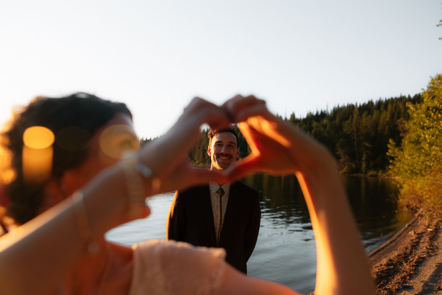 Couple making a heart shape with their hands by a lake at sunset, groom smiling through the heart frame.