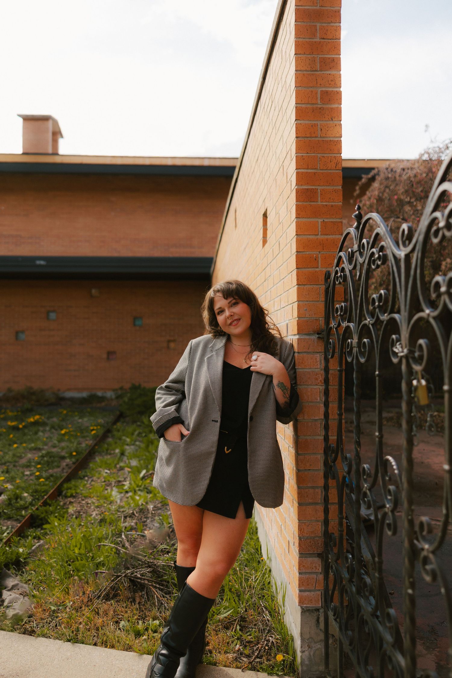 Young woman in gray blazer and black shorts leaning against a brick wall by an ornate iron gate outdoors.