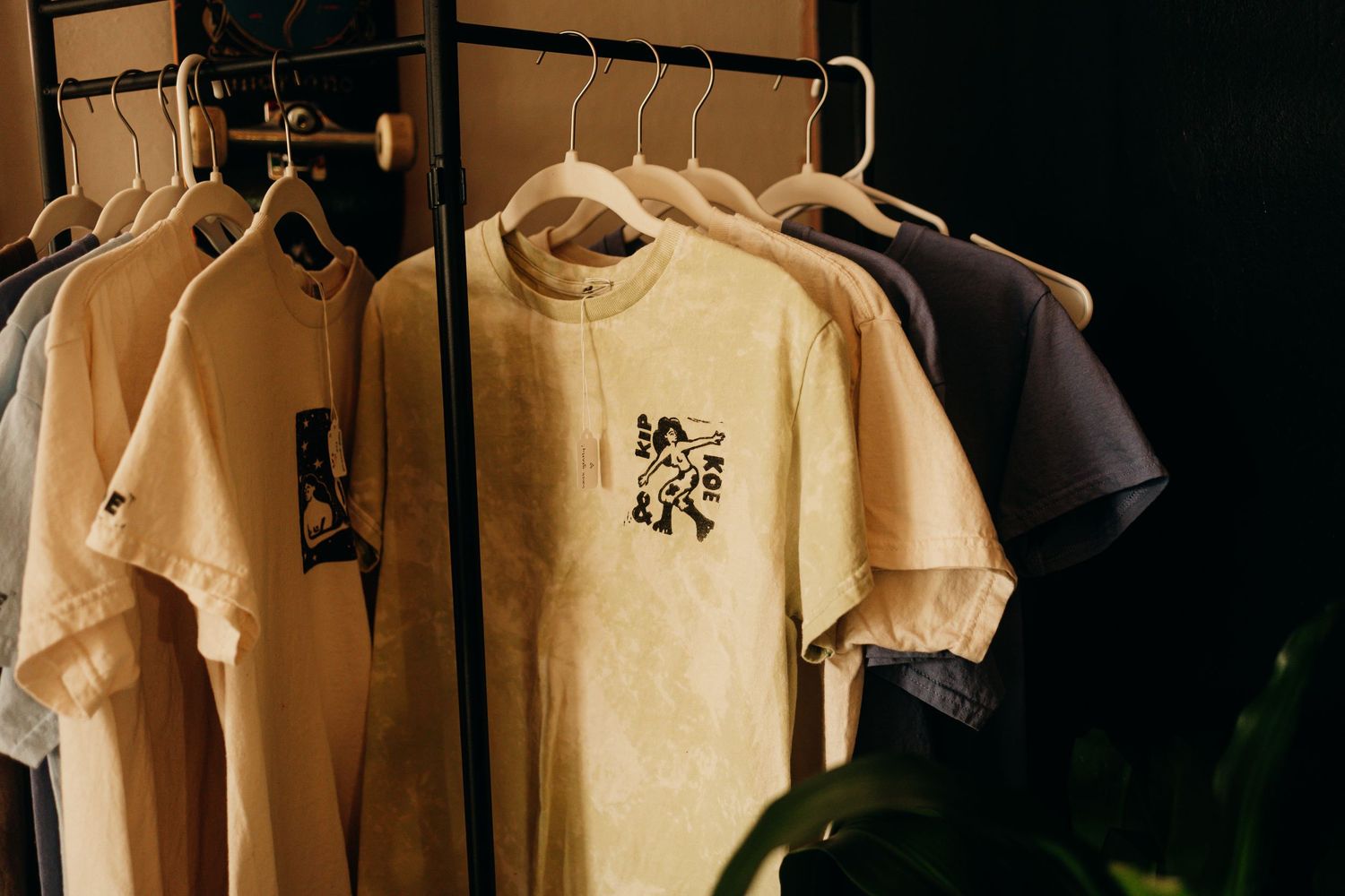 Neutral-toned graphic t-shirts hanging on a black clothing rack in a dark, moody retail display setting.