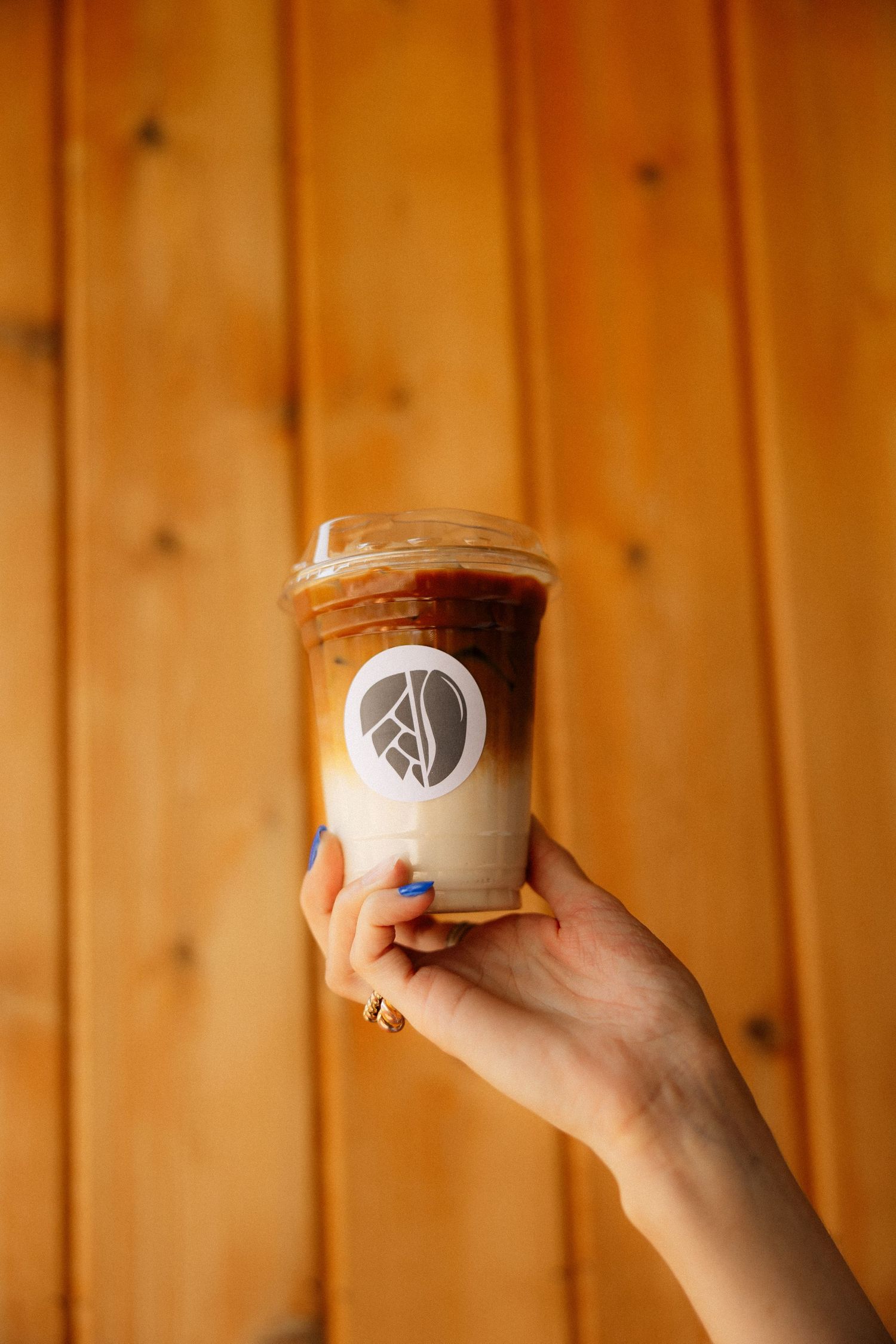 Hand holding an iced coffee in a branded plastic cup against a wooden plank background.