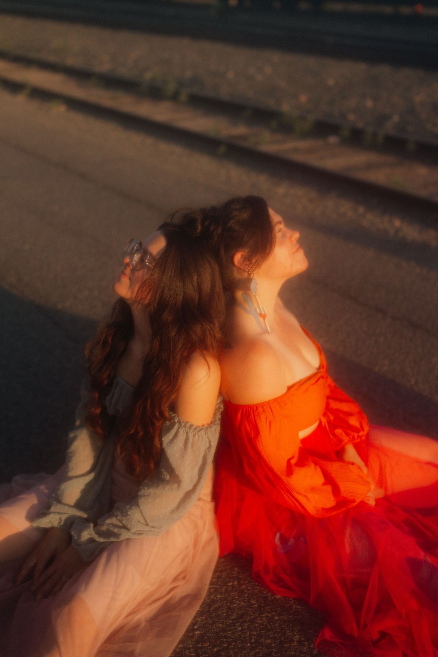 Two women in flowing dresses sitting back-to-back on pavement bathed in warm golden sunset light.