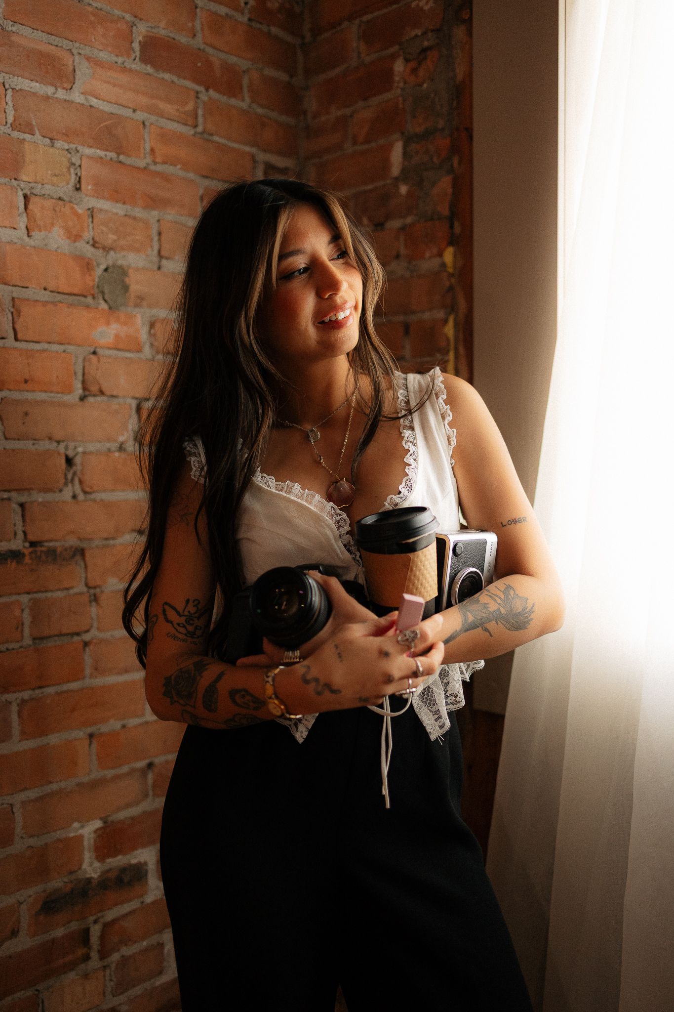 Tattooed woman holding coffee cup and headphones leaning against brick wall near window.