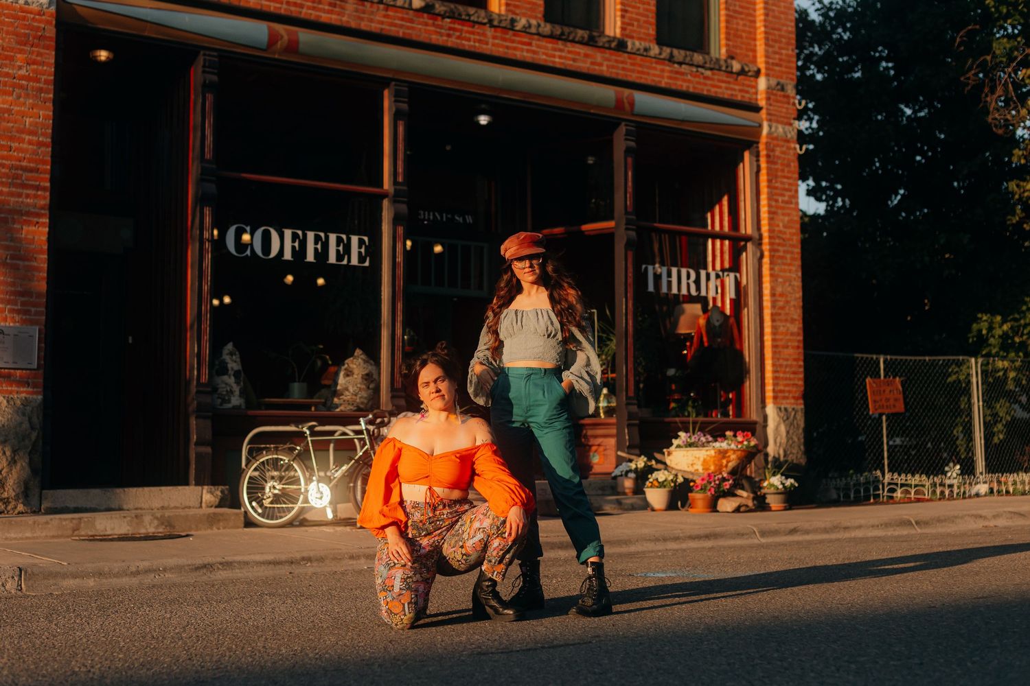 Two stylish women posing outside a Coffee and Thrift shop on a sunny street in vibrant outfits.