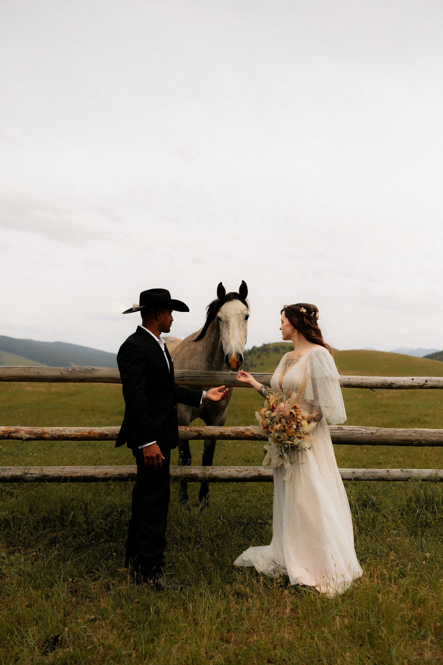 Bride and groom in western attire feed a white horse at a rustic fence with green mountain backdrop.
