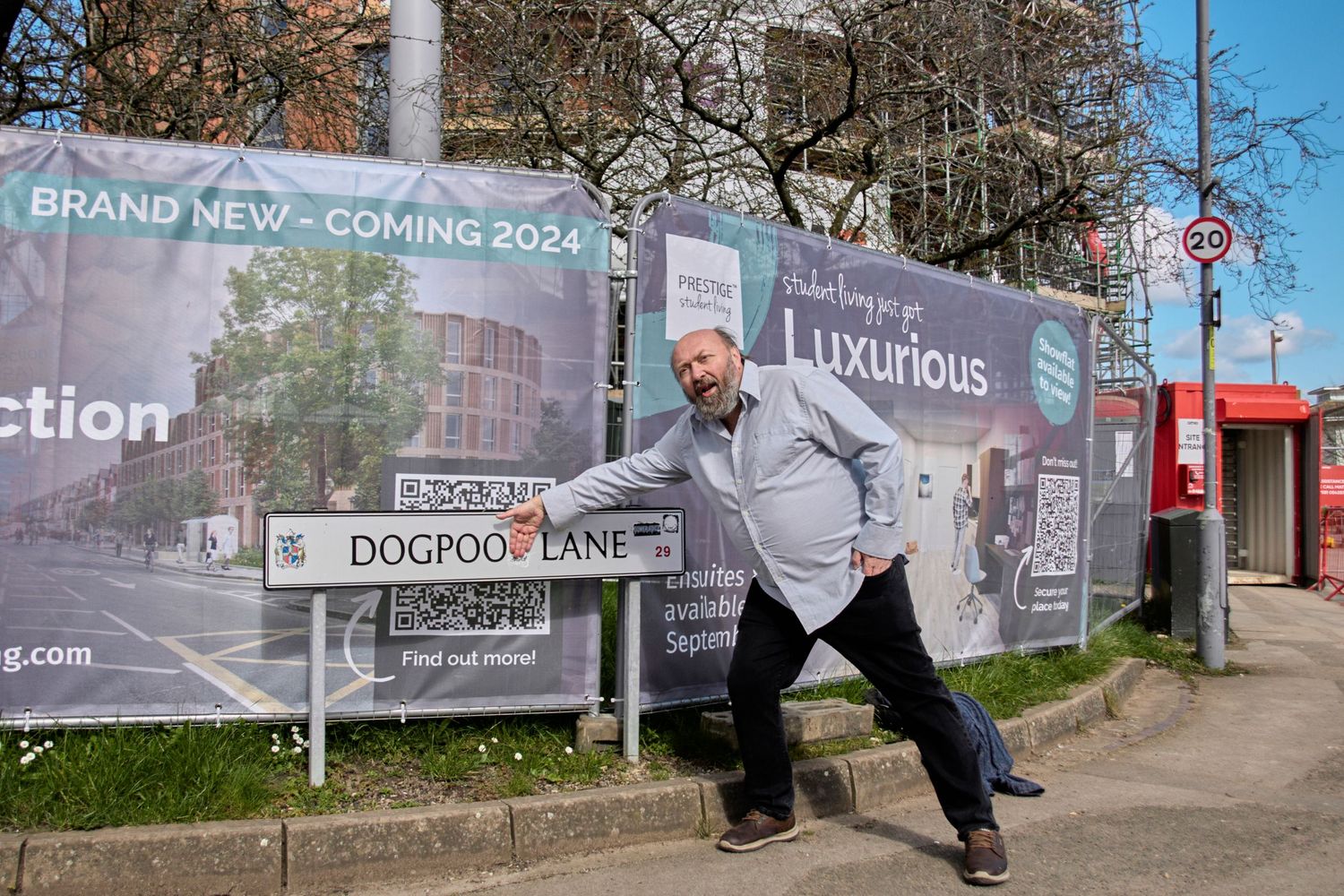 Person posing playfully in front of construction site advertising hoarding on sidewalk.