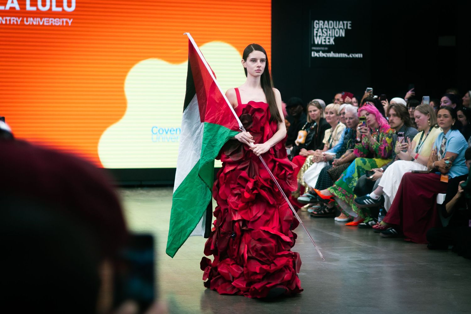 Model in red ruffled gown carries Palestinian flag during fashion show with orange backdrop.