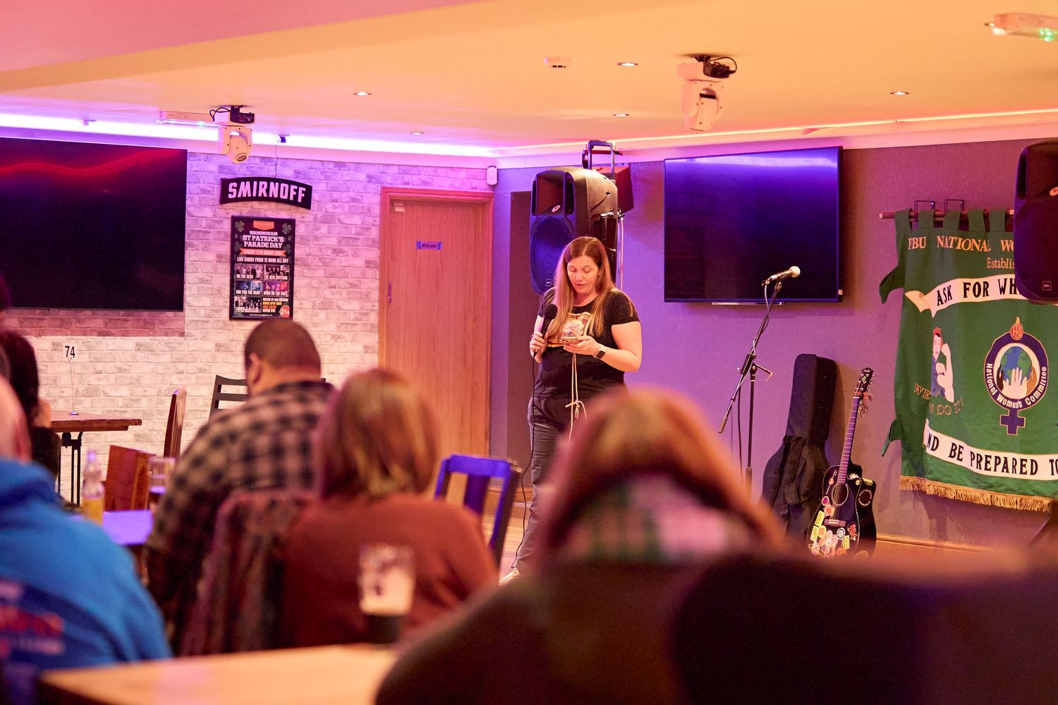 A performer sings on stage in a small venue with purple lighting and an audience watching from tables.