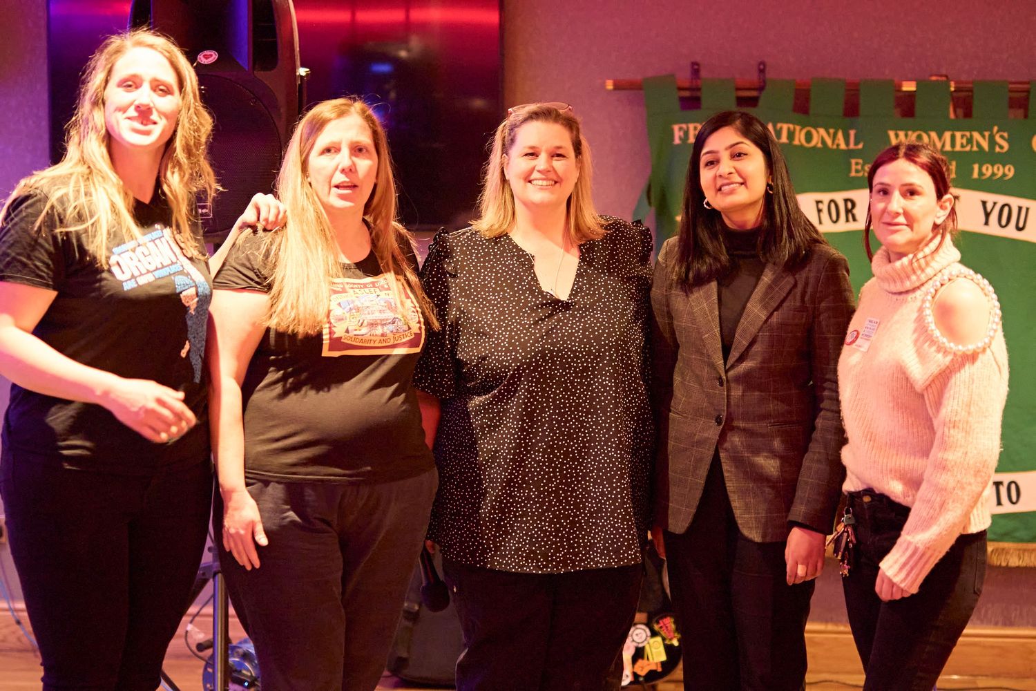 Group of performers posing and smiling together on stage with red lighting in the background.
