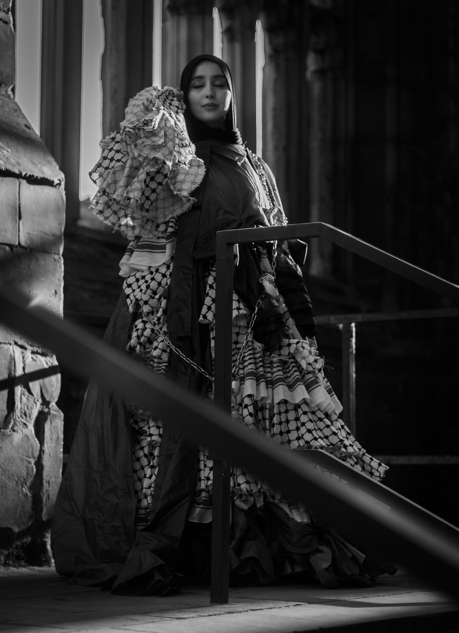 A black and white fashion portrait featuring a dramatic ruffled checkered dress on stairs in moody lighting.