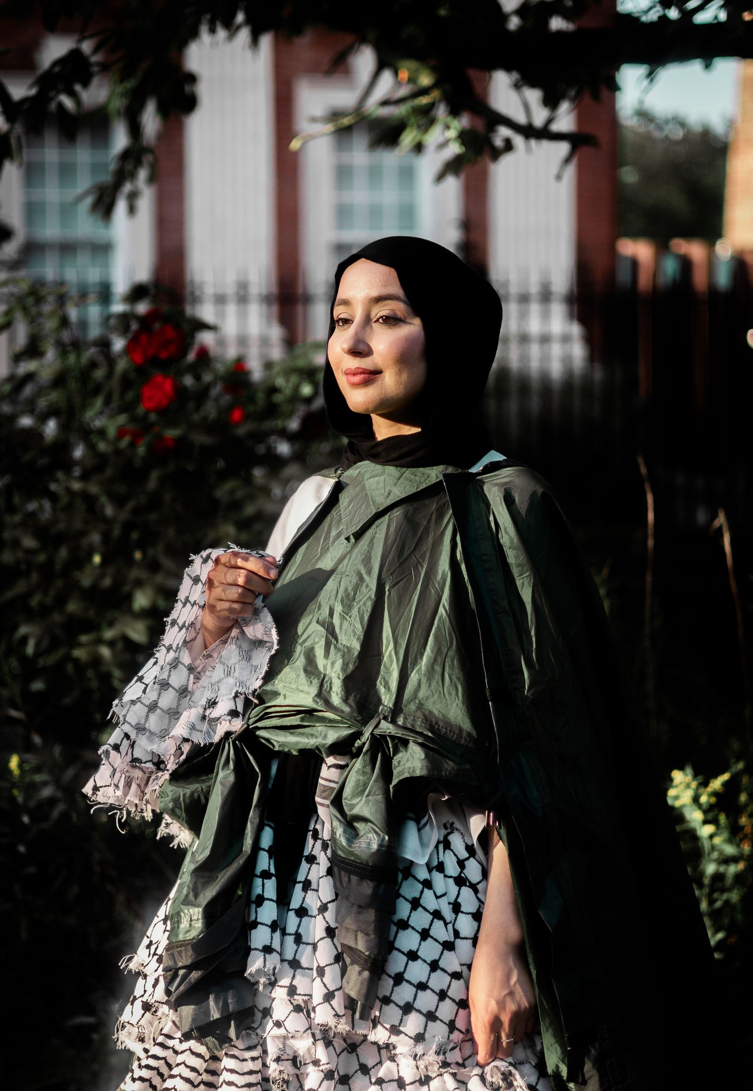 Stylish figure in olive green jacket and black hijab poses with water bottle against lush garden background at sunset.